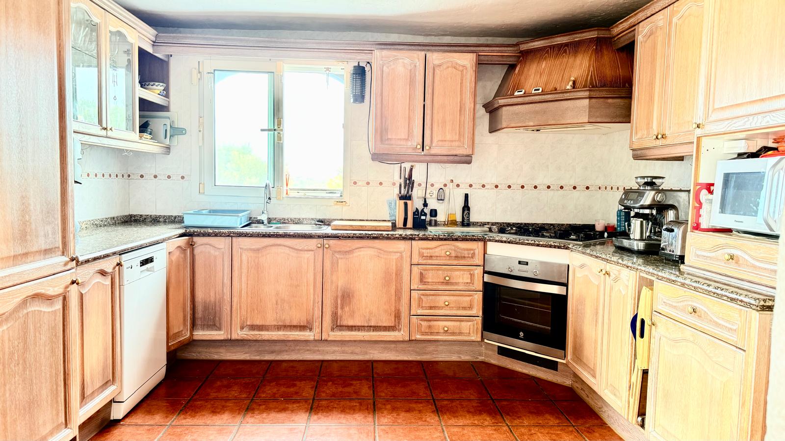 Cocina de estilo tradicional con mobiliario de roble, encimeras de granito, suelo de terracota y una ventana que aporta luz natural sobre la zona del fregadero.