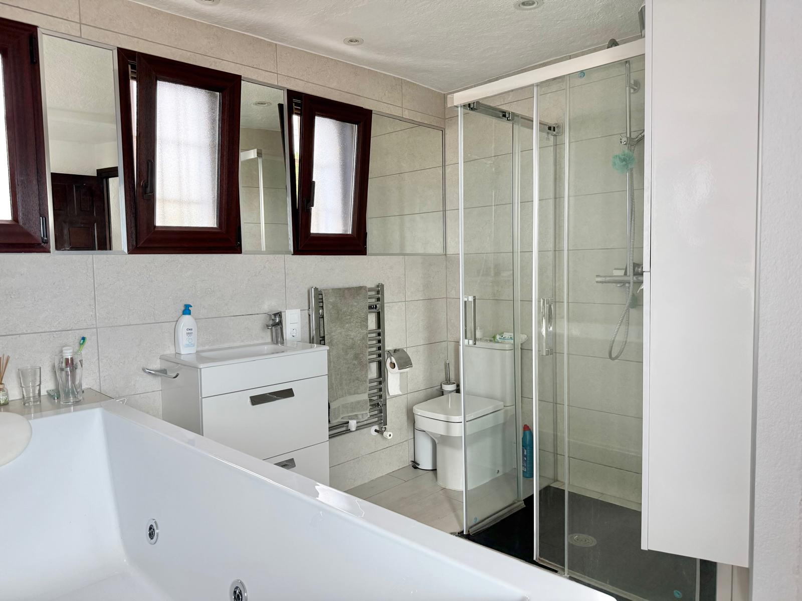 Cuarto de baño completo con bañera de hidromasaje, ducha con mampara de cristal, mueble lavabo blanco y radiador toallero sobre paredes de gres porcelánico.