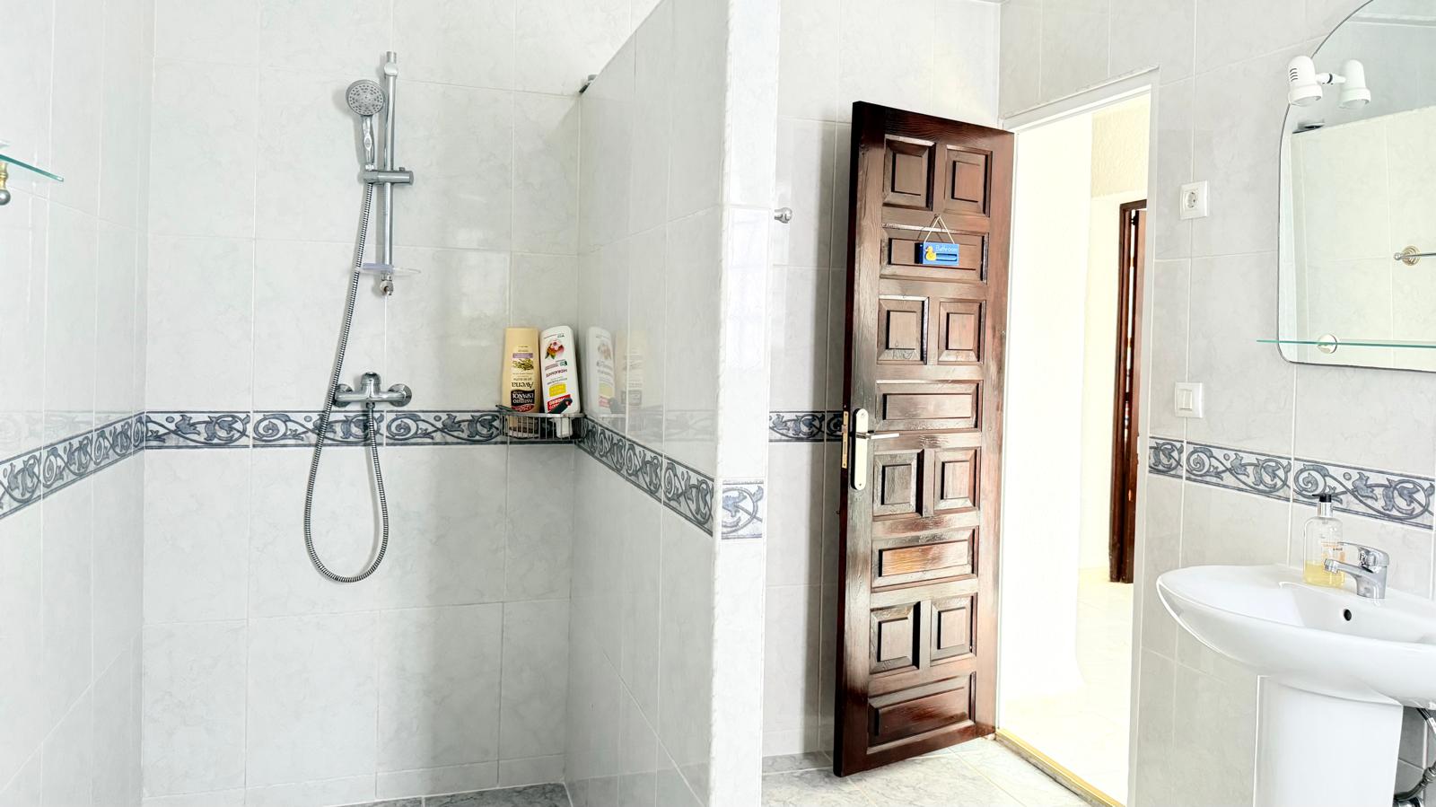 Cuarto de baño con ducha abierta revestida en azulejos blancos con cenefa decorativa, lavabo de pedestal y puerta de madera maciza con paneles tallados.