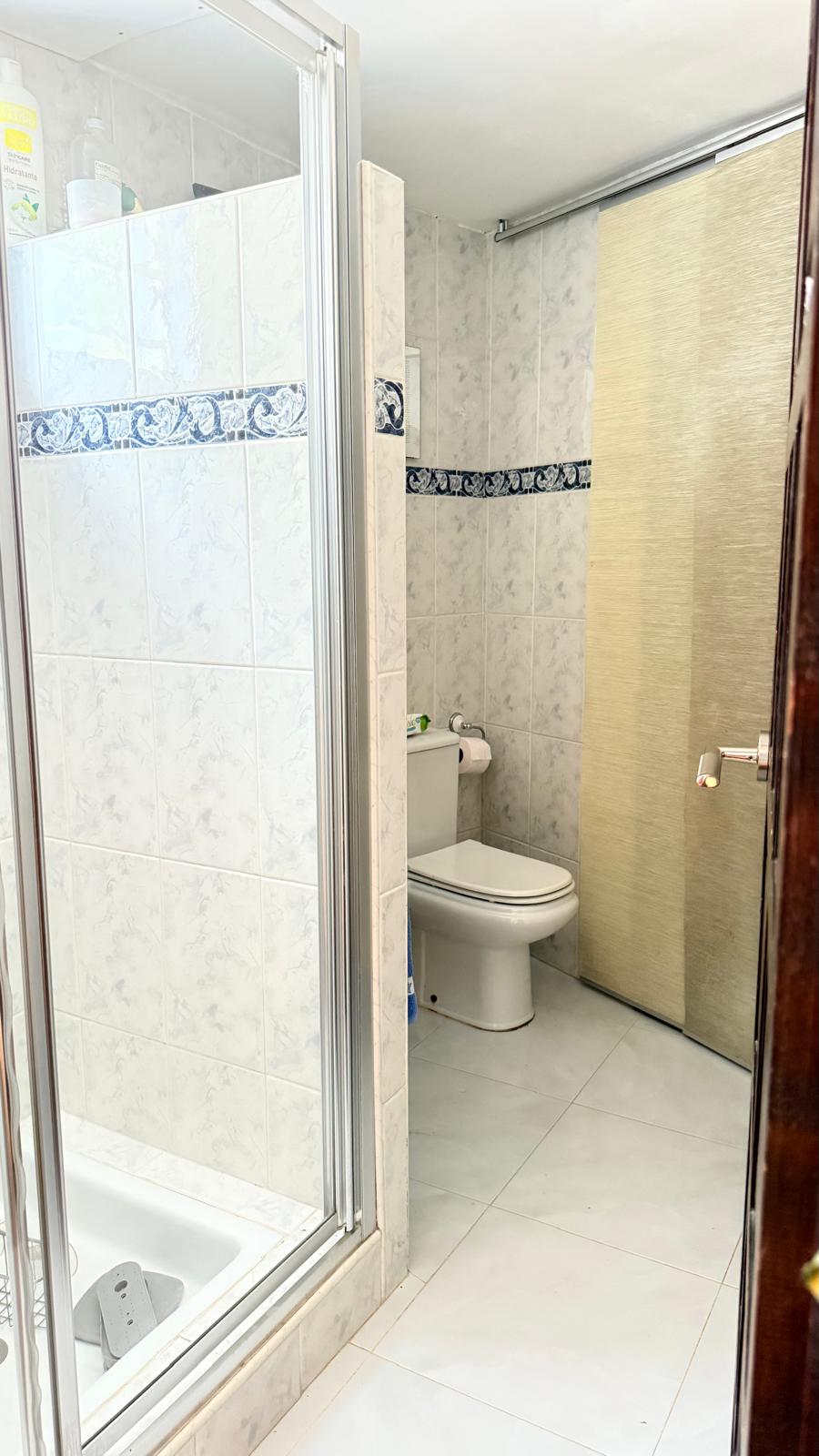 Cuarto de baño con ducha acristalada, azulejos blancos decorados con cenefa azul y un inodoro de porcelana blanca. Incluye una puerta corredera de acabado textil.