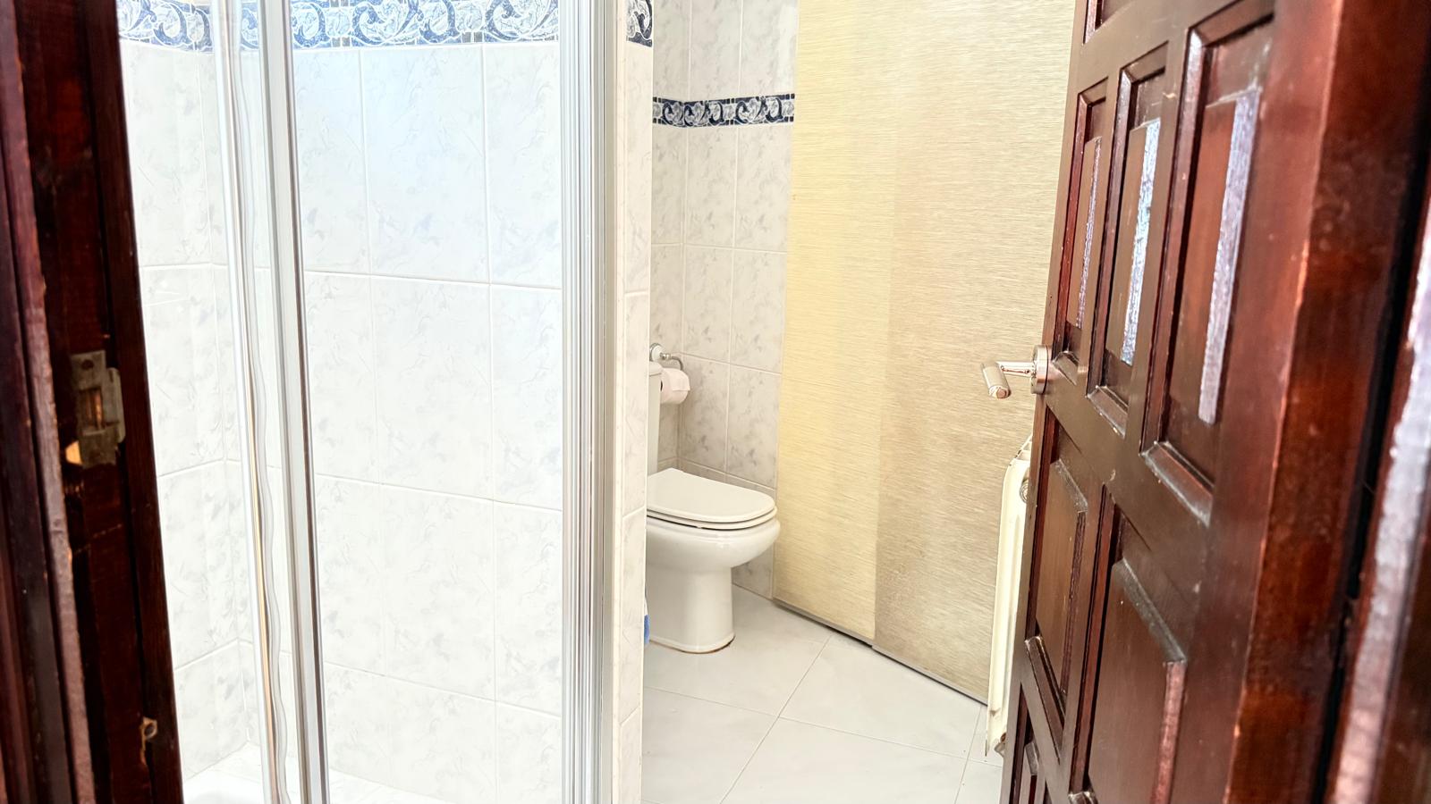 Cuarto de baño con ducha acristalada, inodoro y paredes alicatadas con cenefa decorativa azul. Incluye una puerta de madera oscura en el acceso.