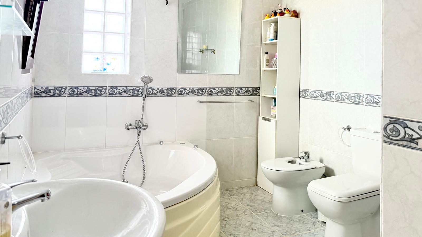 Cuarto de baño equipado con una amplia bañera de hidromasaje en esquina, bidet e inodoro, con paredes de azulejo blanco y una cenefa decorativa clásica.
