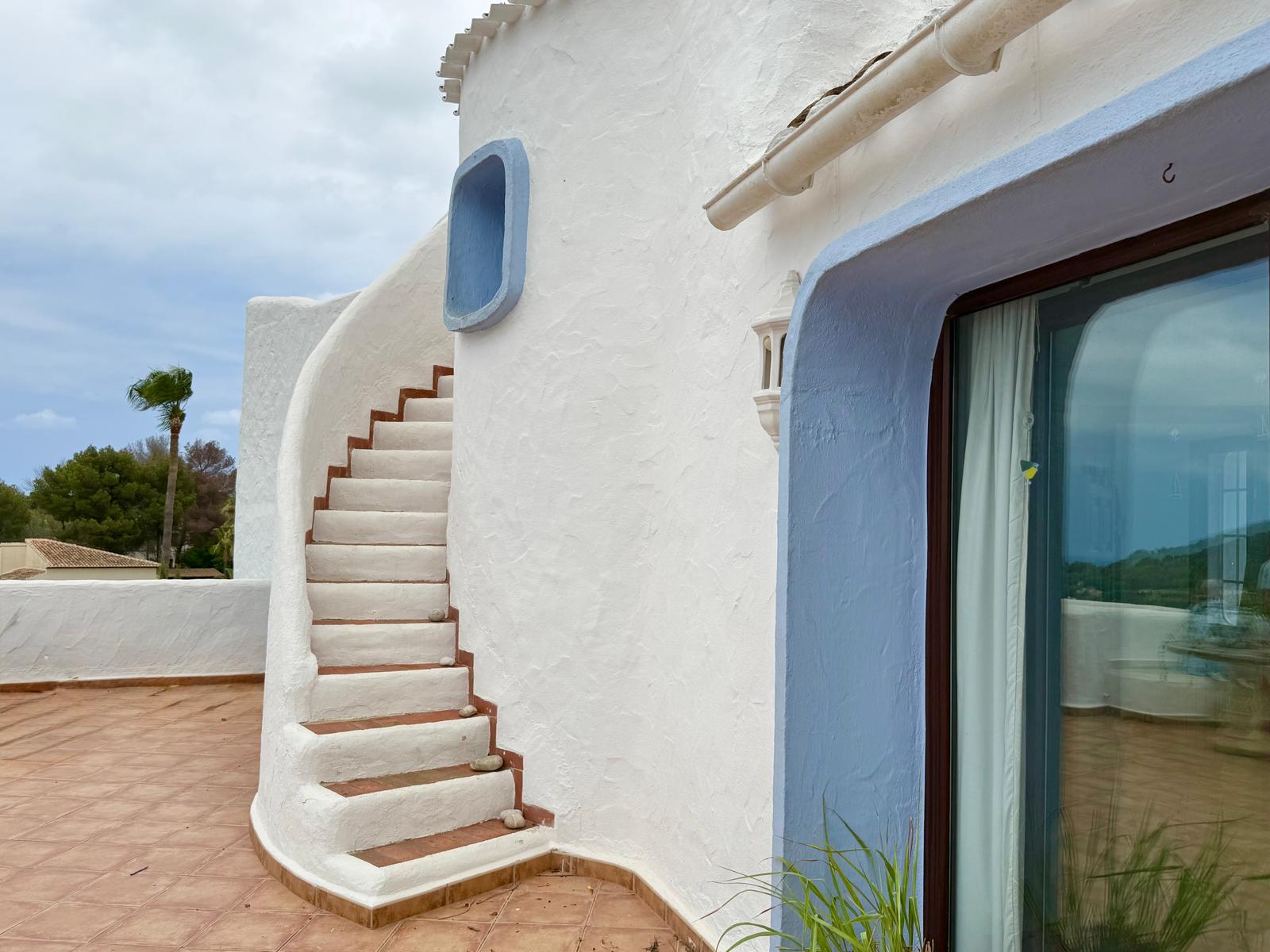 Detalle de la fachada blanca de estilo ibicenco con escalera exterior curva, peldaños de barro cocido y marcos resaltados en azul sobre la terraza.