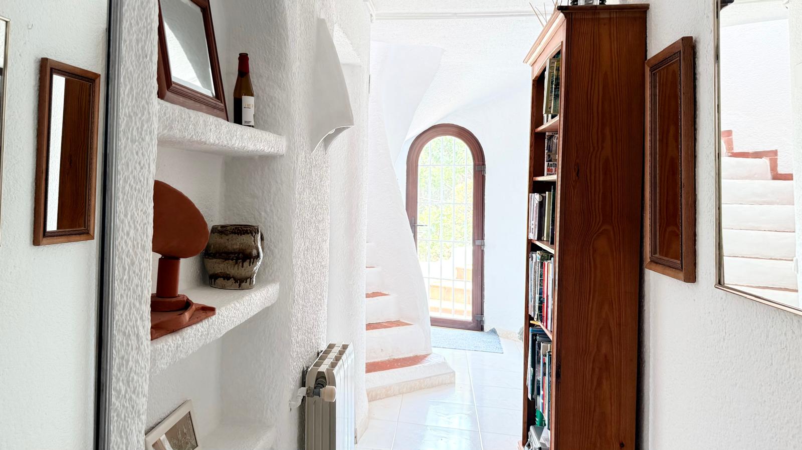 Distribuidor con paredes blancas texturizadas, estanterías de obra y una escalera interior curva que resalta el diseño arquitectónico original.