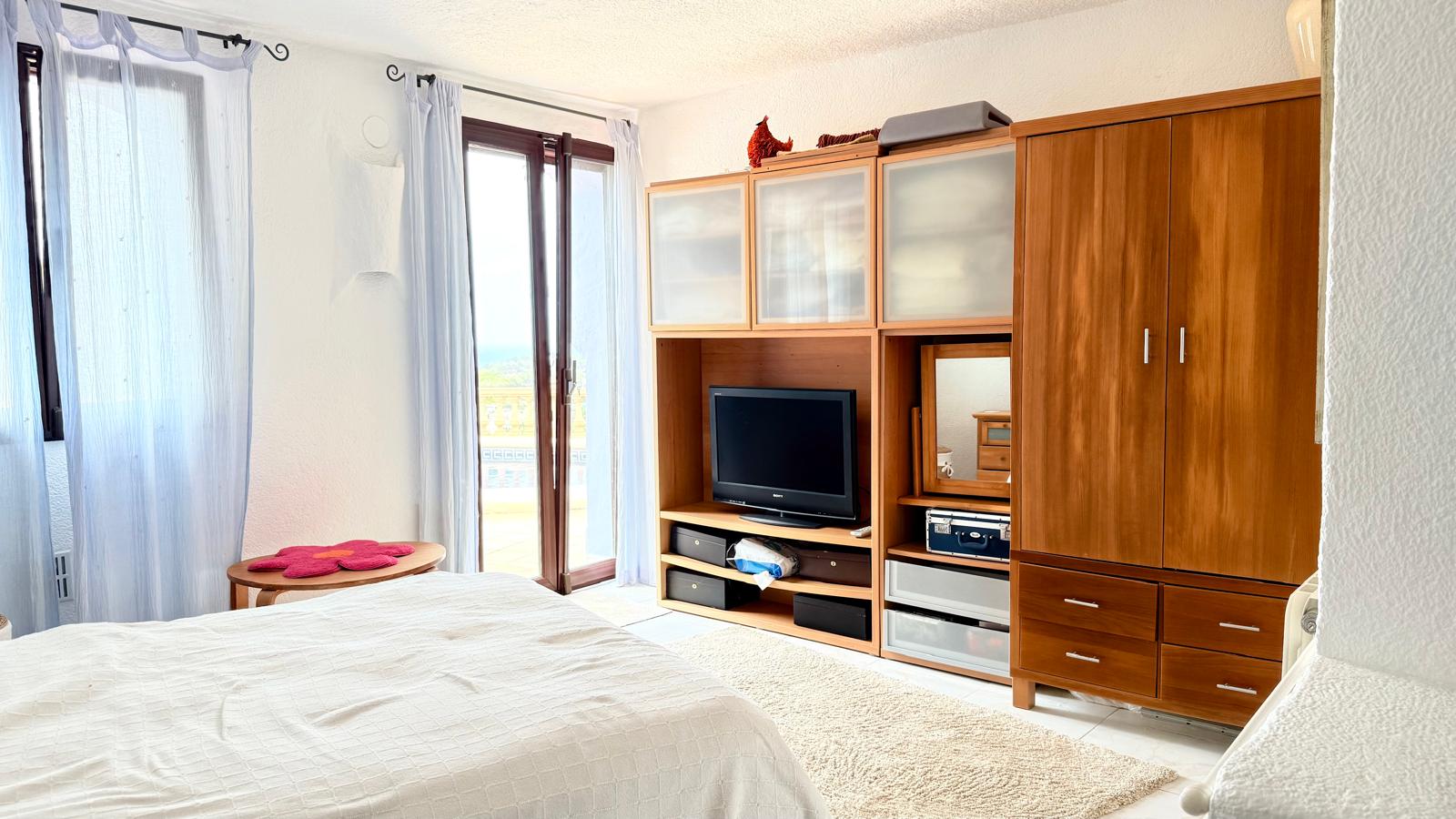 Dormitorio luminoso con mobiliario de madera integrado, zona de TV y acceso directo a la terraza con vistas al mar a través de puertas acristaladas.