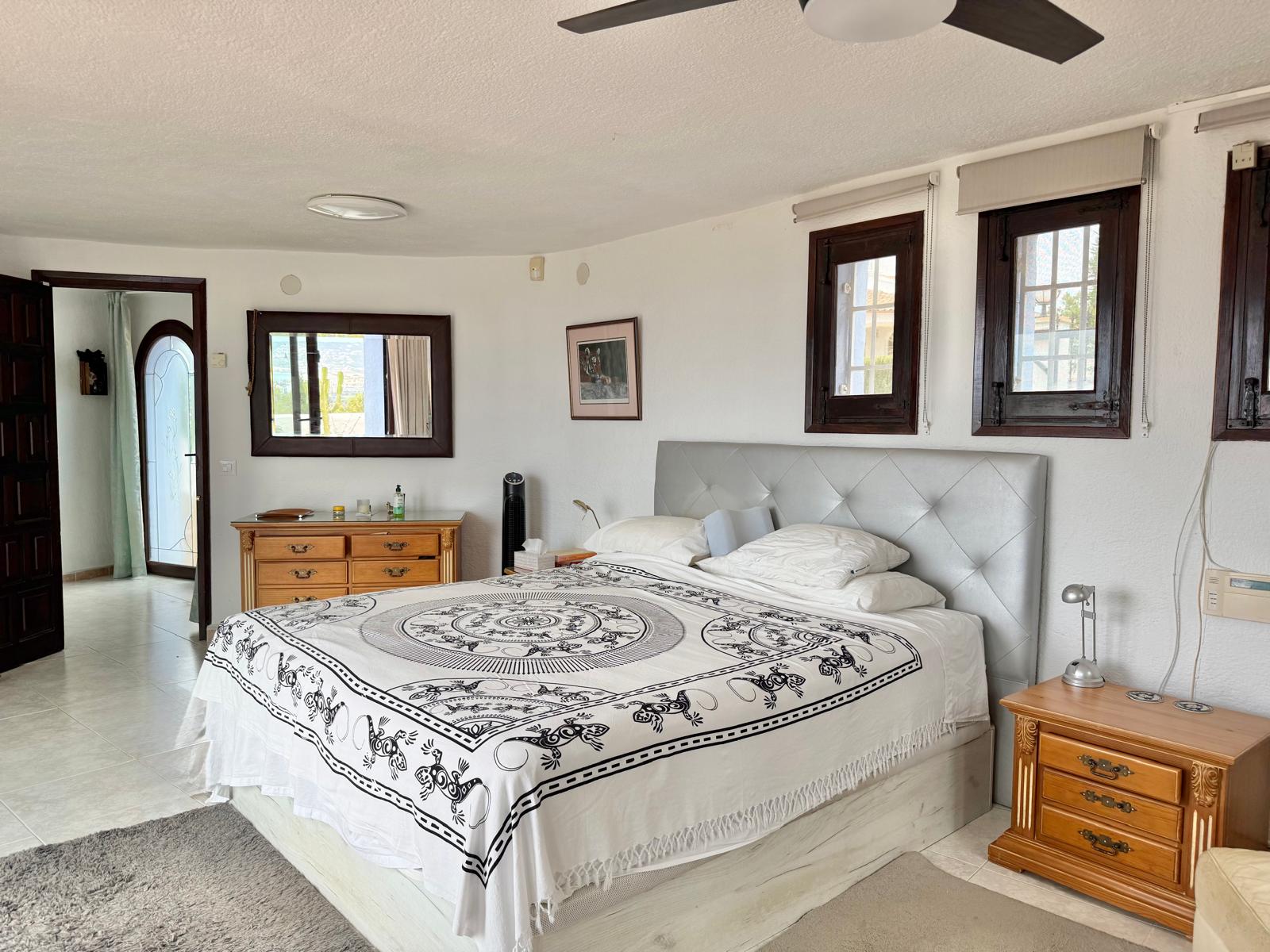 Dormitorio principal con paredes curvas, mobiliario de madera clásica y ventanas de marco oscuro. La estancia dispone de ventilador de techo y suelos cerámicos claros.