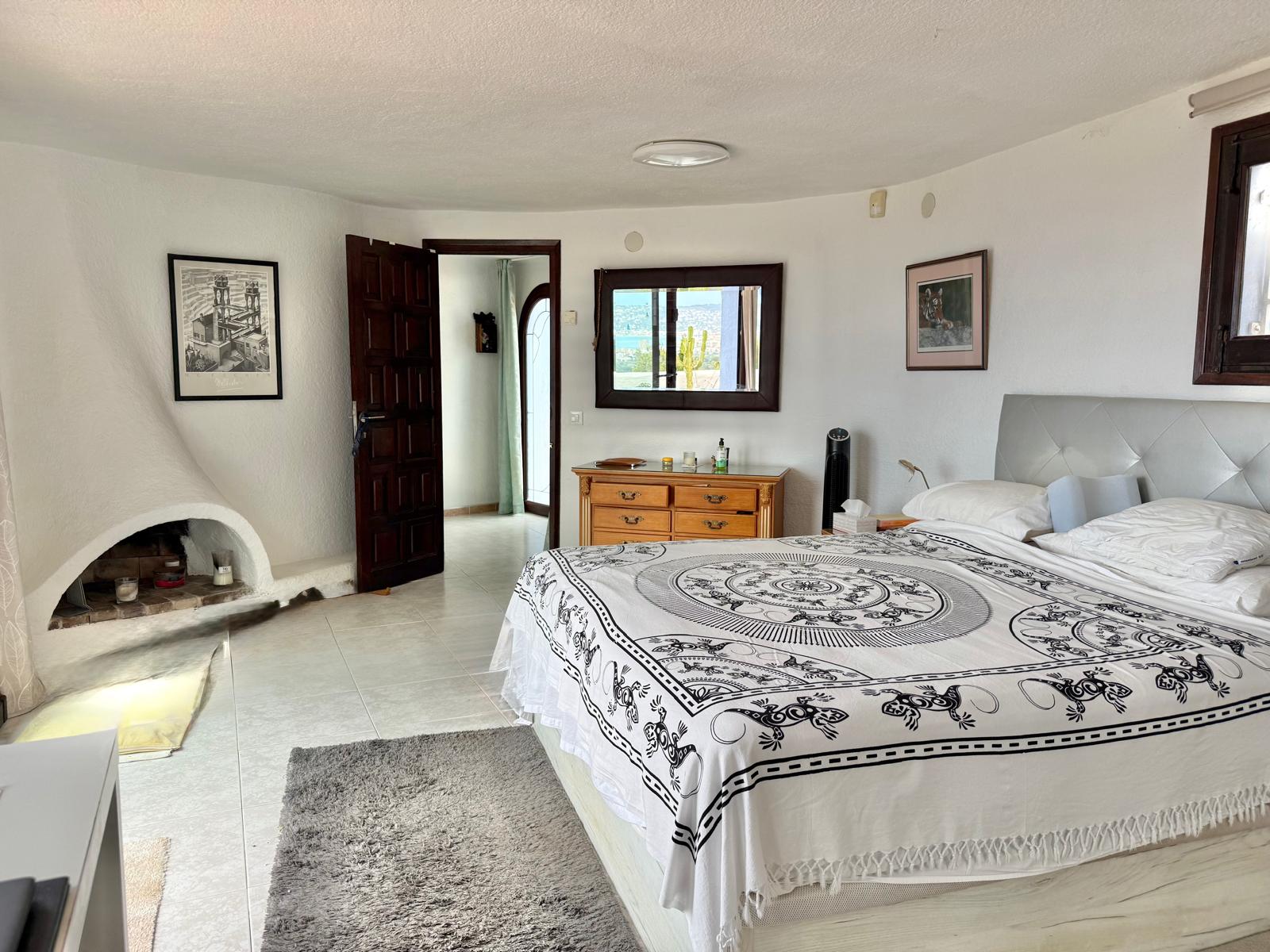 Dormitorio principal de estilo ibicenco con chimenea de obra integrada, paredes blancas texturizadas y suelos cerámicos claros que aportan luminosidad.