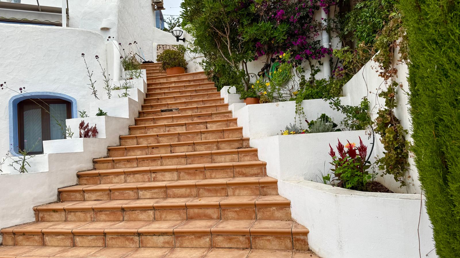 Escalera exterior de baldosas cerámicas que conecta los niveles de la villa, flanqueada por jardineras blancas integradas con vegetación mediterránea y buganvillas.