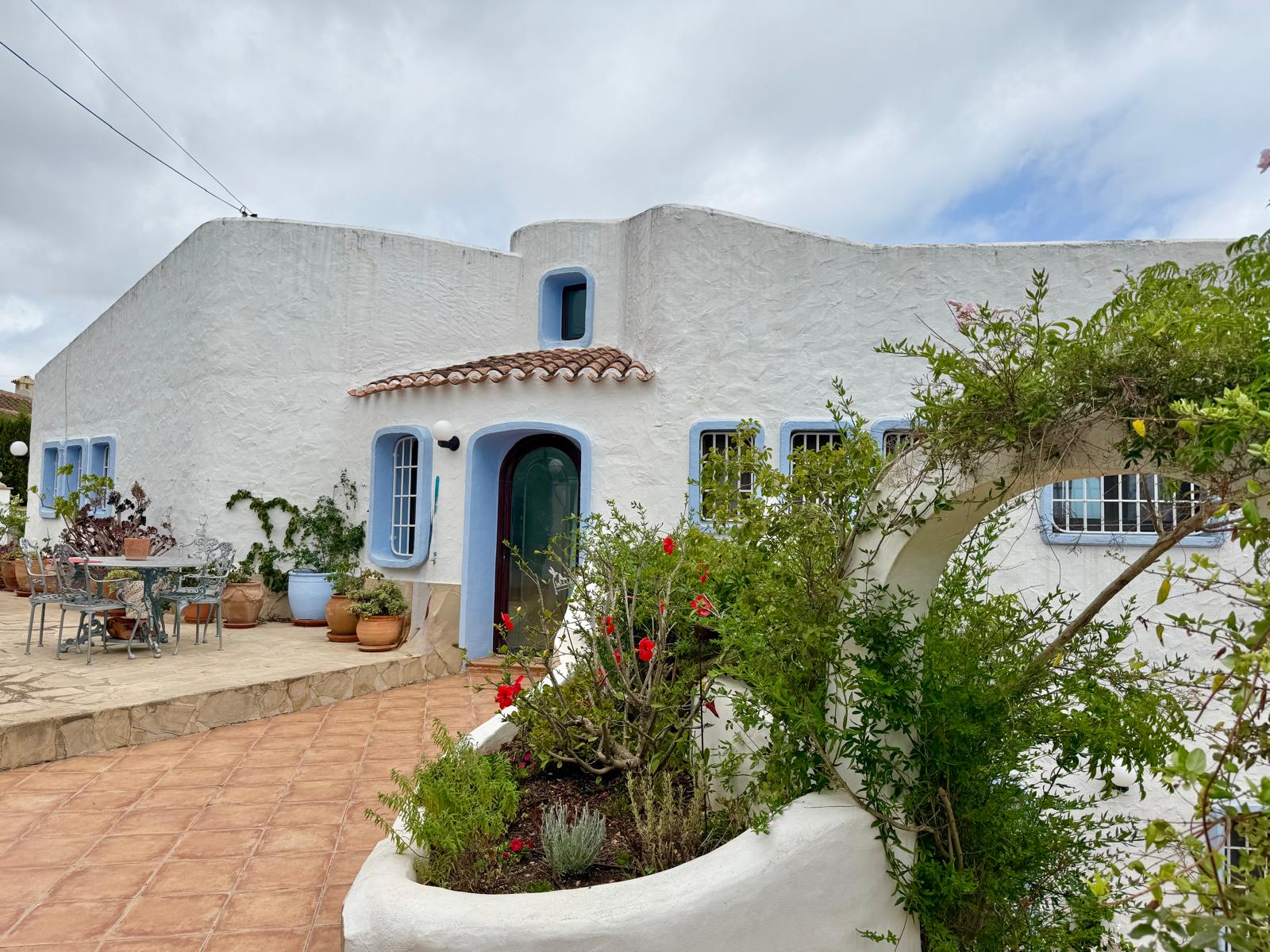 Fachada de estilo ibicenco con muros blancos orgánicos, carpintería azul y terraza de baldosas de barro rodeada de vegetación mediterránea y macetas decorativas.