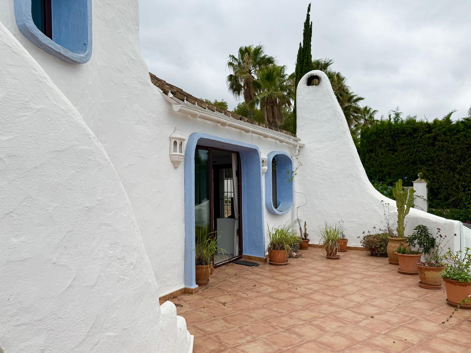 Fachada de estilo ibicenco con muros blancos orgánicos, marcos azules y una chimenea escultural. La terraza de baldosas cerámicas incluye diversas plantas mediterráneas en macetas.