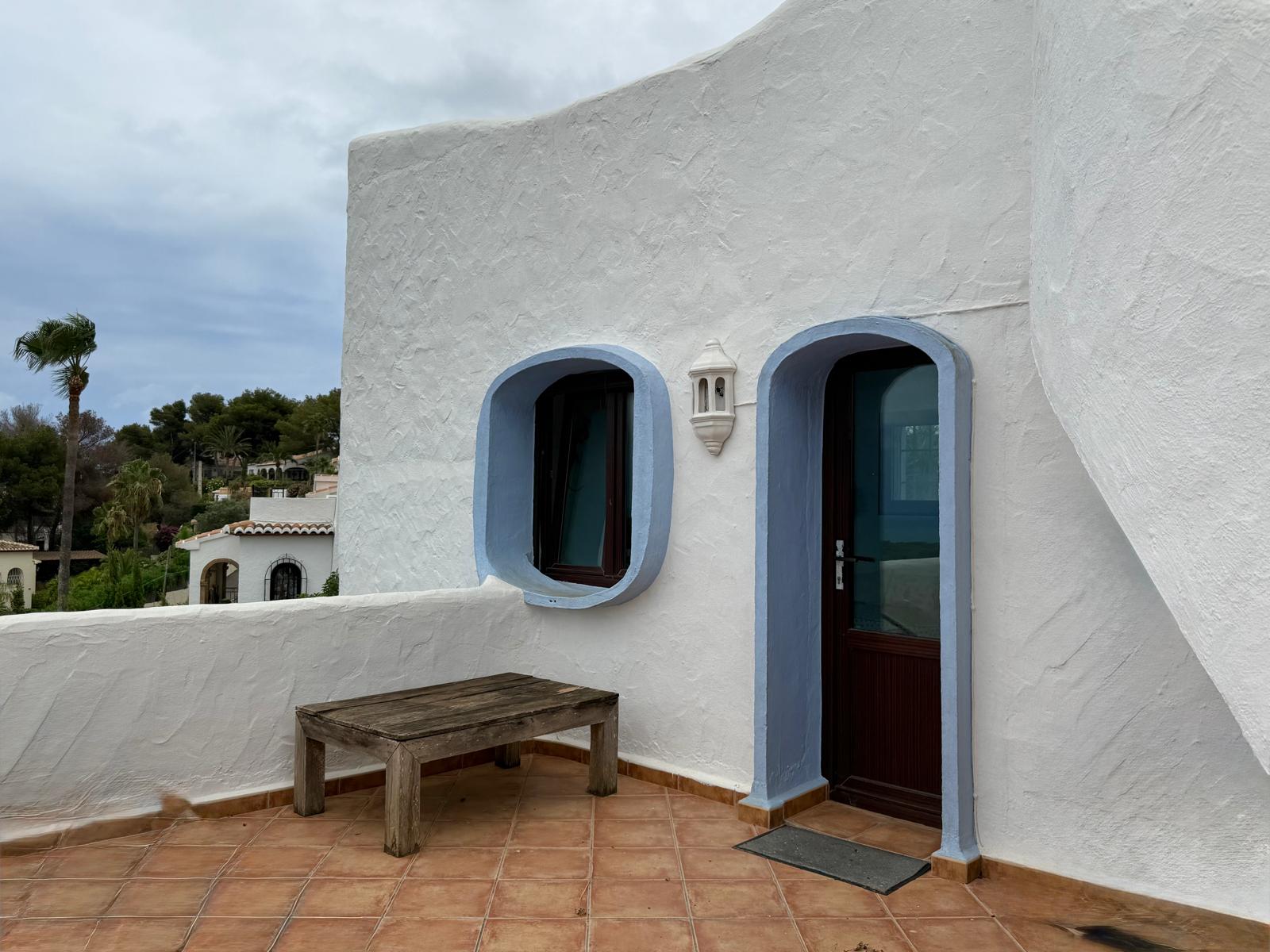 Fachada de estilo ibicenco con muros blancos texturizados, marcos arqueados en azul y suelos de terracota. Destaca la carpintería de madera oscura y el mobiliario rústico de la terraza.
