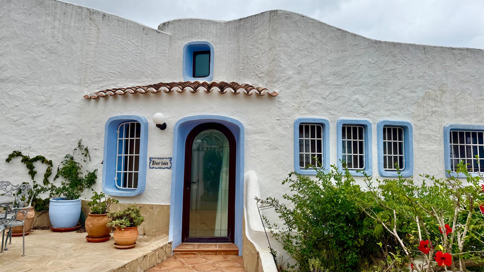 Fachada de estilo ibicenco con muros blancos texturizados, marcos azules y puerta arqueada. La entrada se complementa con macetas de terracota y vegetación mediterránea.