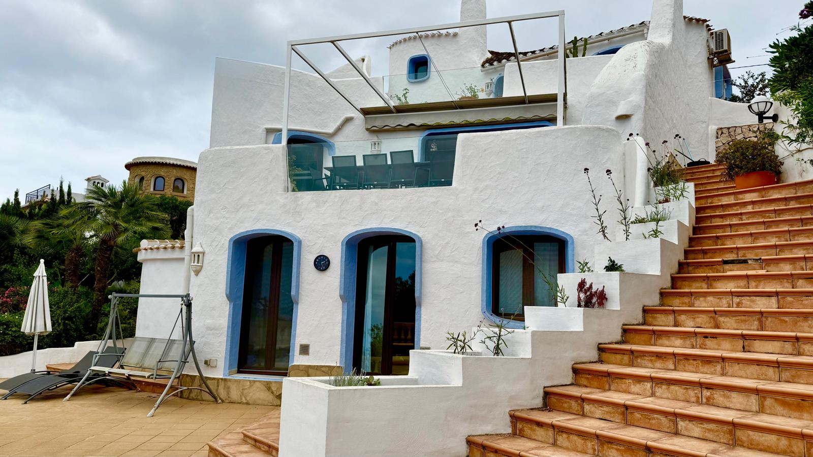 Fachada de estilo ibicenco con muros blancos y detalles azules. Se aprecian los niveles de la villa conectados por escaleras de terracota y una terraza superior con barandilla de cristal.