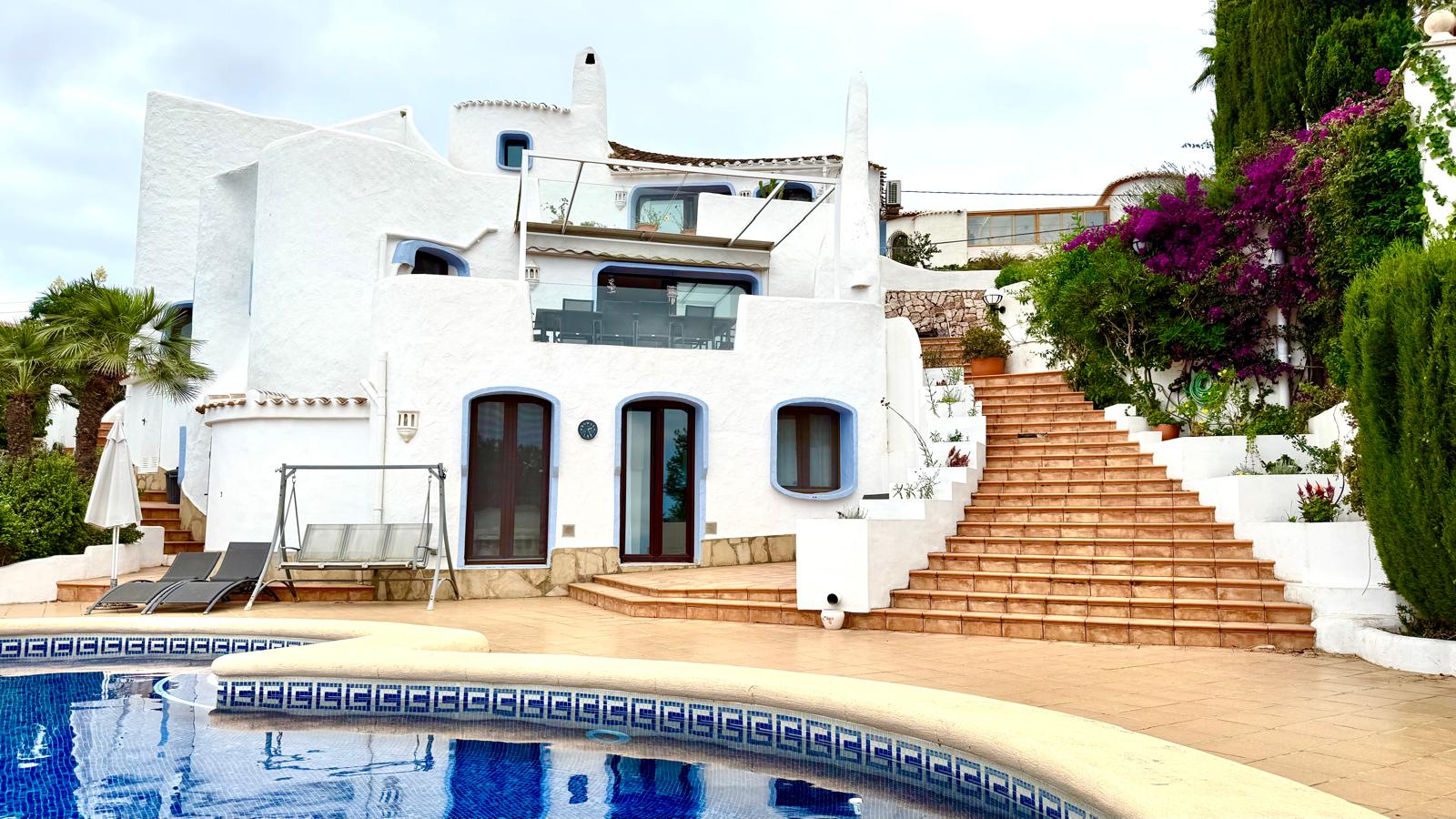 Fachada de estilo ibicenco con muros blancos y detalles azules, terraza de baldosas cerámicas, amplia escalera exterior y piscina rodeada de vegetación mediterránea.