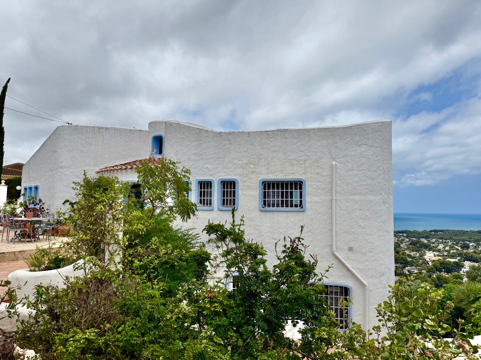 Fachada de estilo ibicenco con muros curvos blancos y detalles en azul, situada en una posición elevada con vistas panorámicas a la costa de Jávea.