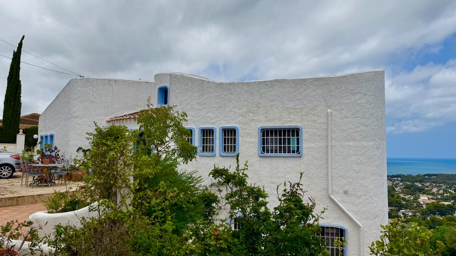 Fachada de estilo ibicenco con paredes blancas texturizadas, carpintería azul y ventanas protegidas. La terraza exterior ofrece vistas abiertas hacia la costa de Jávea.