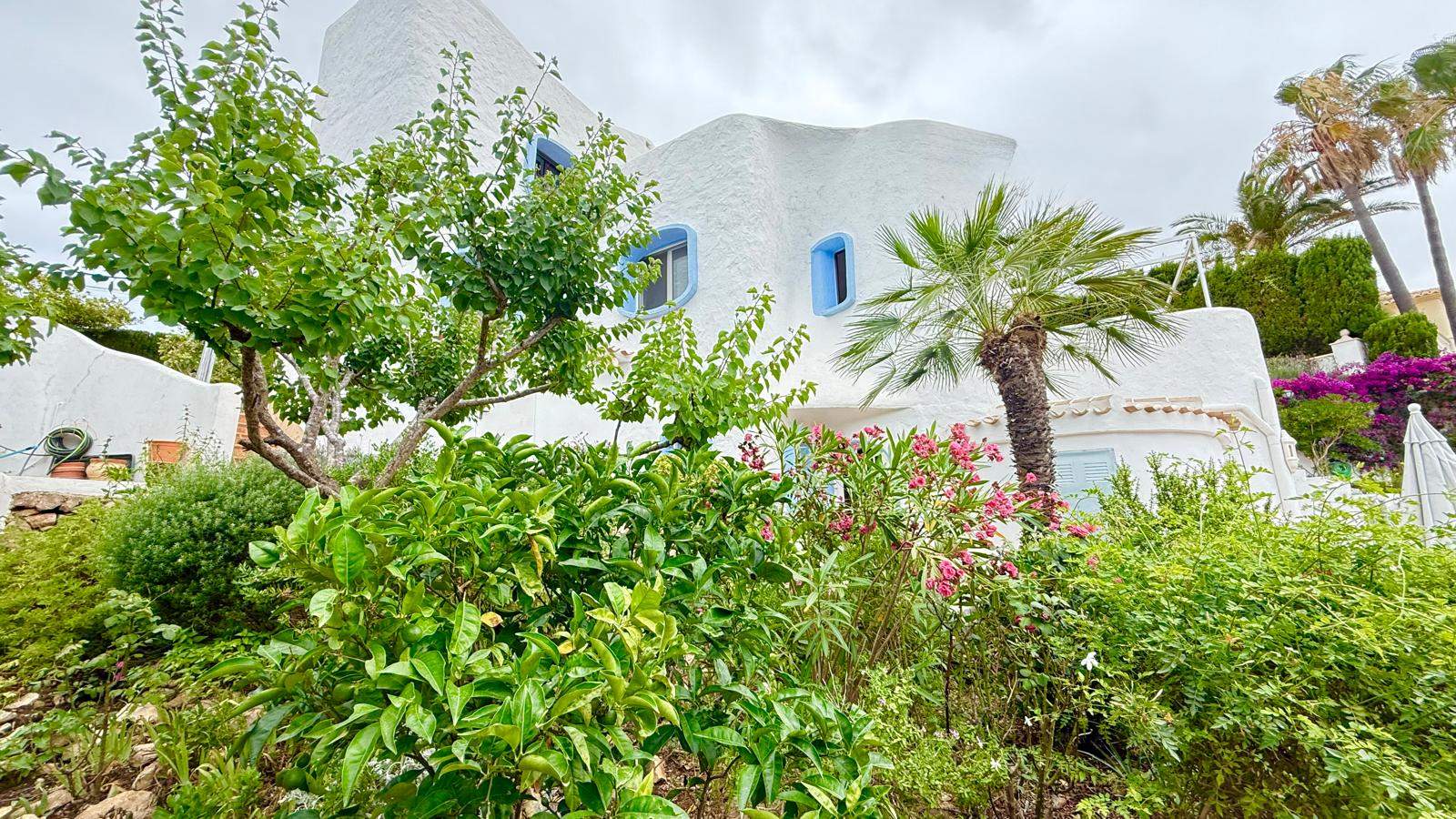 Fachada de estilo ibicenco con paredes blancas texturizadas y ventanas de marcos azules, rodeada de un jardín mediterráneo con palmeras y vegetación autóctona.