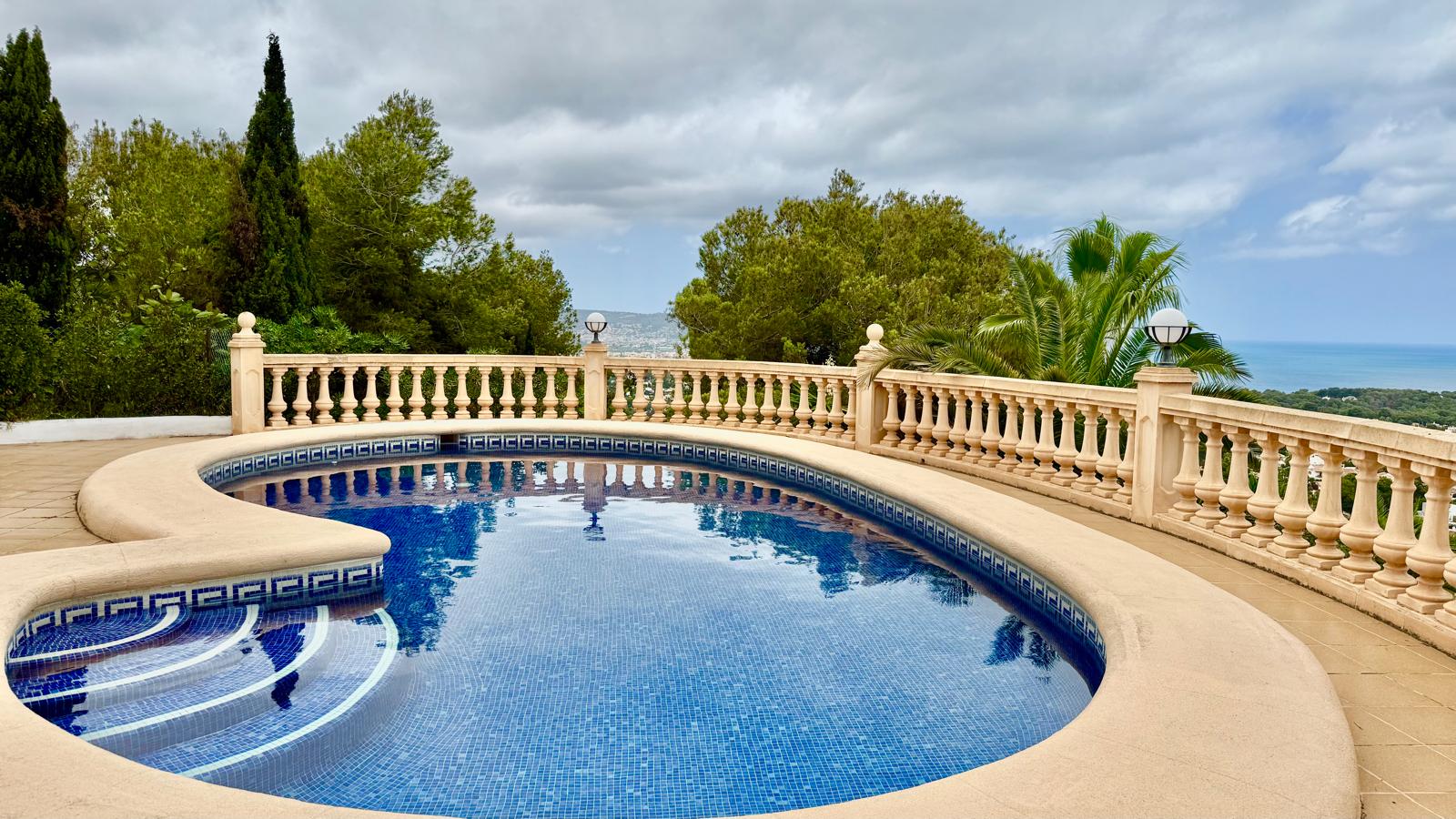 Piscina privada de diseño curvo con gresite azul, rodeada por una terraza pavimentada y balaustrada clásica con vistas panorámicas al mar y entorno arbolado.