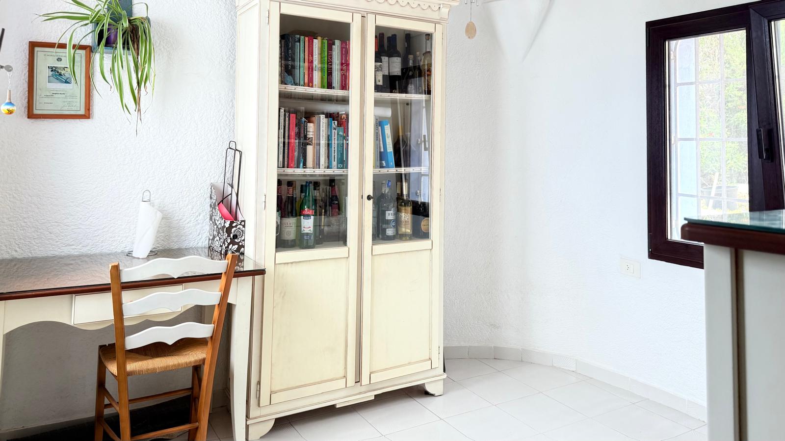 Rincón de trabajo con escritorio de madera blanca, silla rústica y una vitrina clásica que alberga libros y botellas sobre un suelo de baldosas claras.