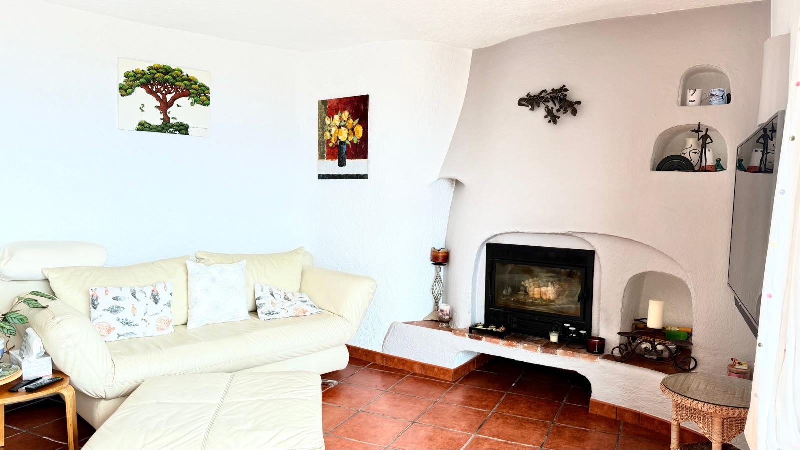 Salón de estilo ibicenco con chimenea de cassette integrada en una estructura curva con hornacinas decorativas, suelos de terracota y paredes blancas texturizadas.