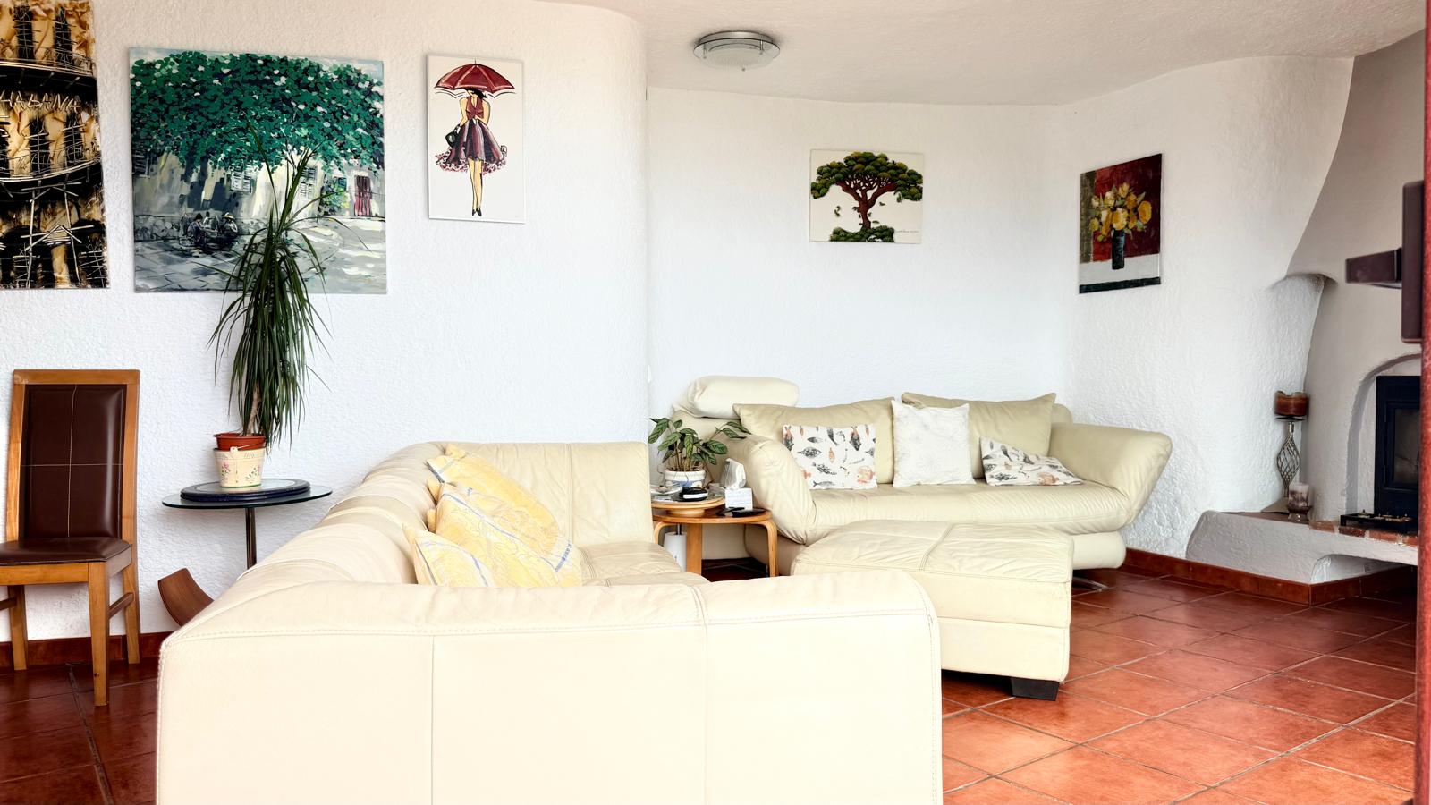 Salón de estilo ibicenco con suelos de terracota, paredes blancas texturizadas y chimenea de leña. Incluye sofás de cuero en tono crema y cuadros decorativos.