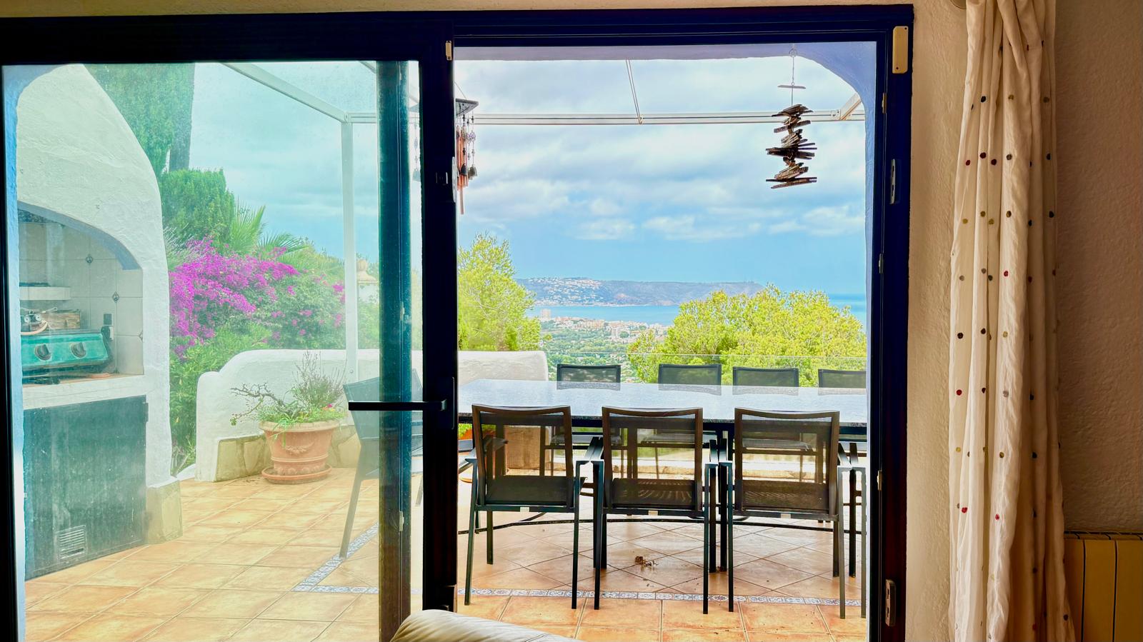 Vista desde el salón hacia la terraza principal con comedor exterior, barbacoa de obra y vistas panorámicas a la bahía de Jávea a través de grandes ventanales.