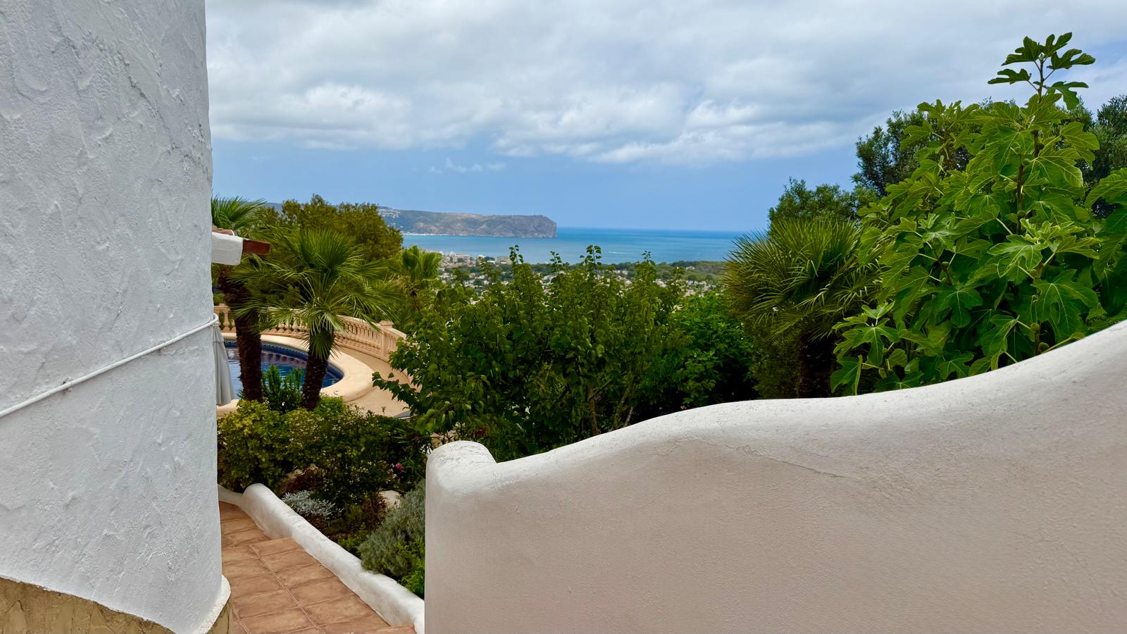 Vista panorámica al mar y la bahía de Jávea desde las escaleras exteriores, rodeadas de muros blancos curvos de estilo ibicenco y vegetación mediterránea madura.