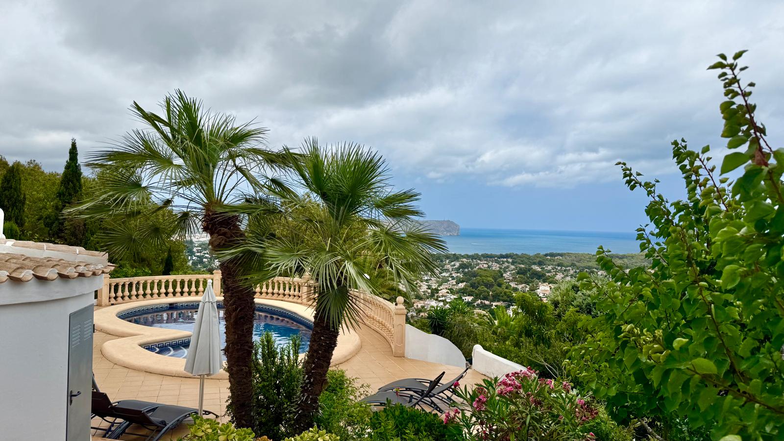 Vista panorámica de la piscina curva y el jardín mediterráneo hacia la bahía de Jávea, con palmeras maduras y balaustrada de piedra sobre el litoral.