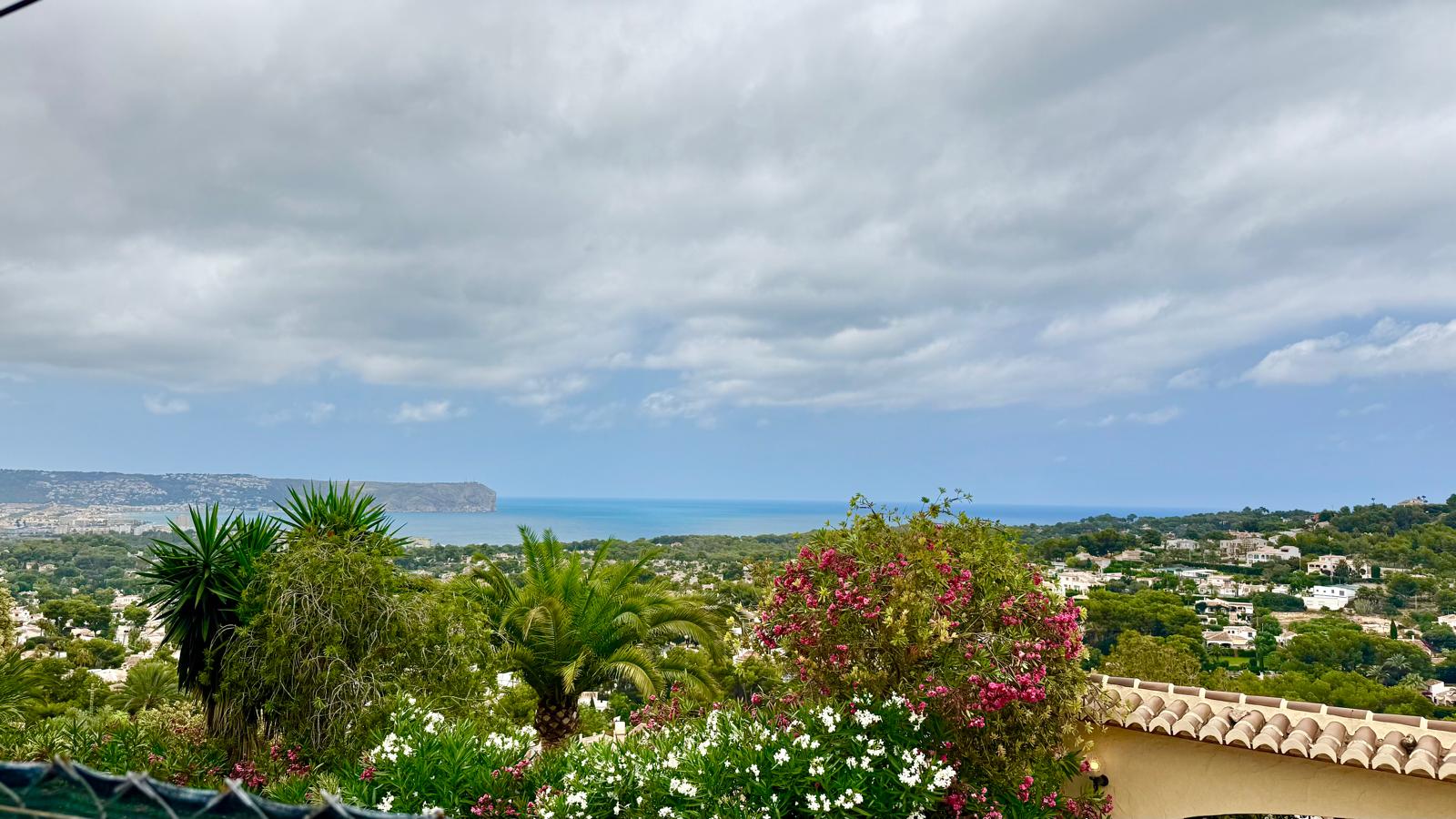 Vista panorámica desde la propiedad hacia la bahía de Jávea y el Cabo de San Antonio, con un jardín mediterráneo de adelfas y palmeras en primer plano.
