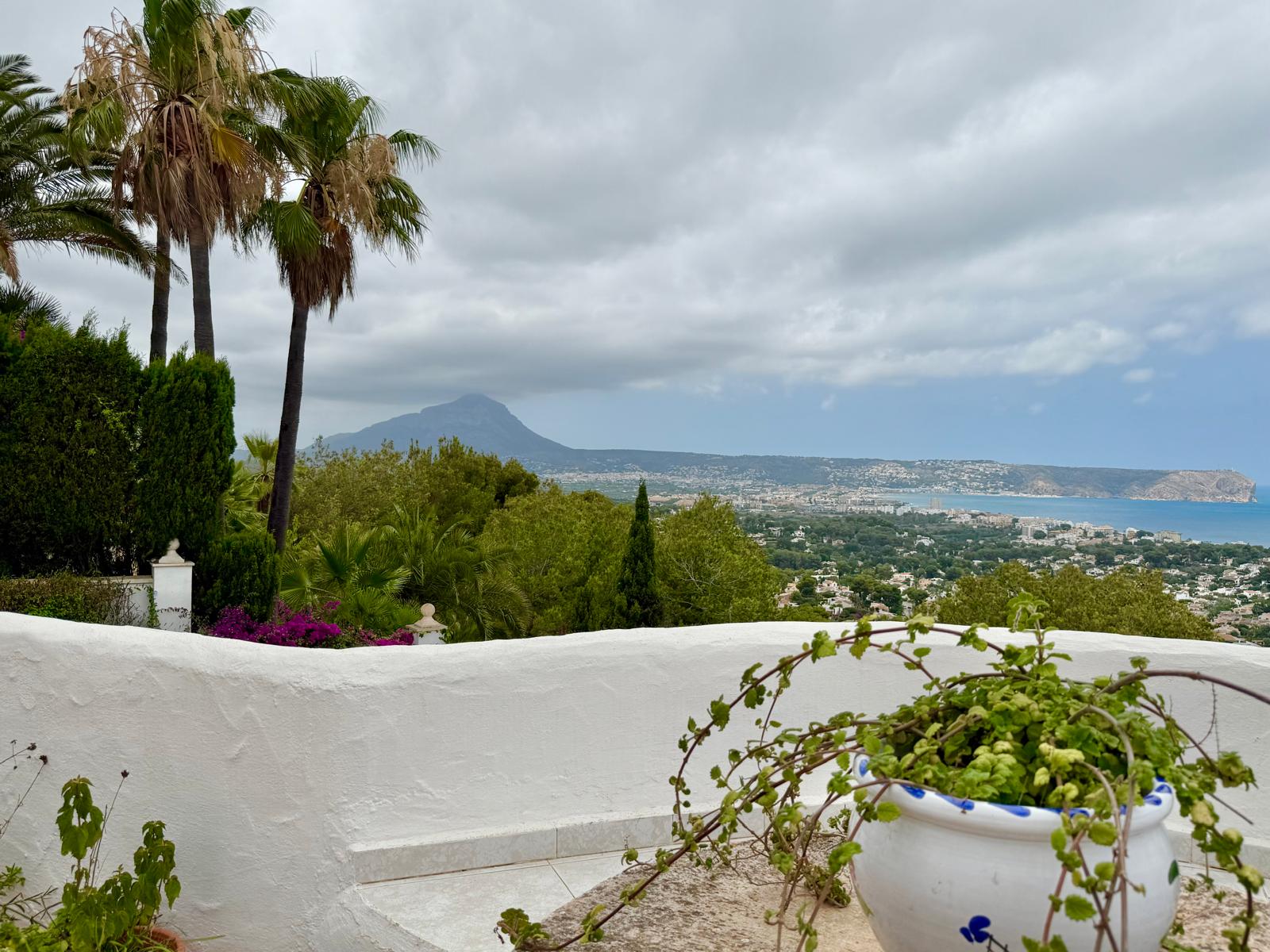 Vista panorámica desde la terraza superior hacia la bahía de Jávea y el Montgó, enmarcada por muros blancos de estilo ibicenco y vegetación mediterránea madura.
