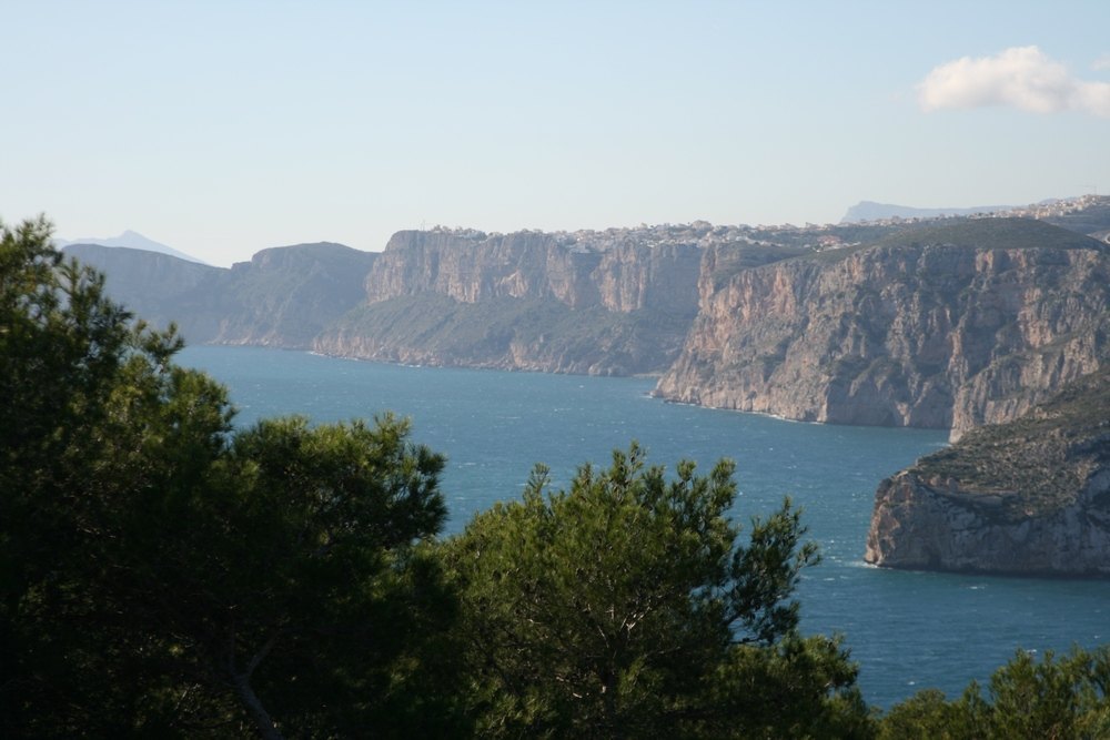 Vistas panorámicas al mar Mediterráneo y a los acantilados de la costa de Jávea, enmarcadas por la vegetación autóctona de pinos bajo un cielo despejado.
