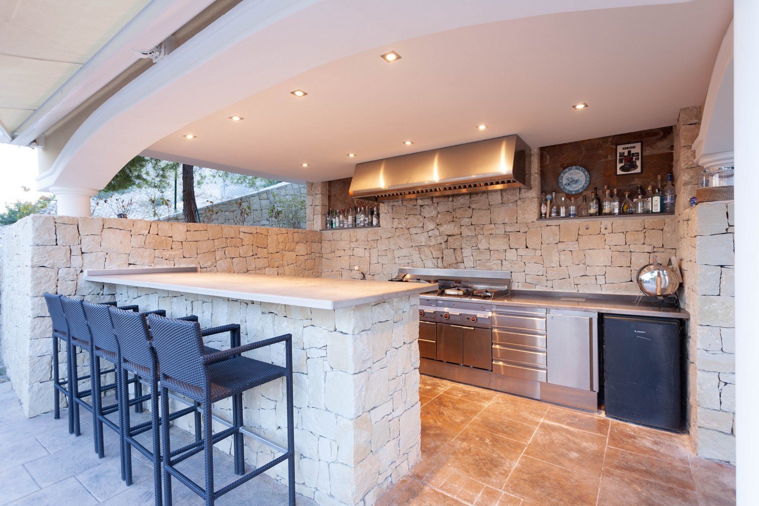 Cocina exterior con barra de piedra, electrodomésticos de acero inoxidable y asientos de ratán.