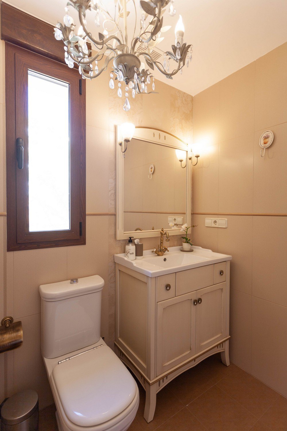 Detalle de baño clásico con lavabo de madera y encimera blanca, espejo con apliques y lámpara de araña de cristal.