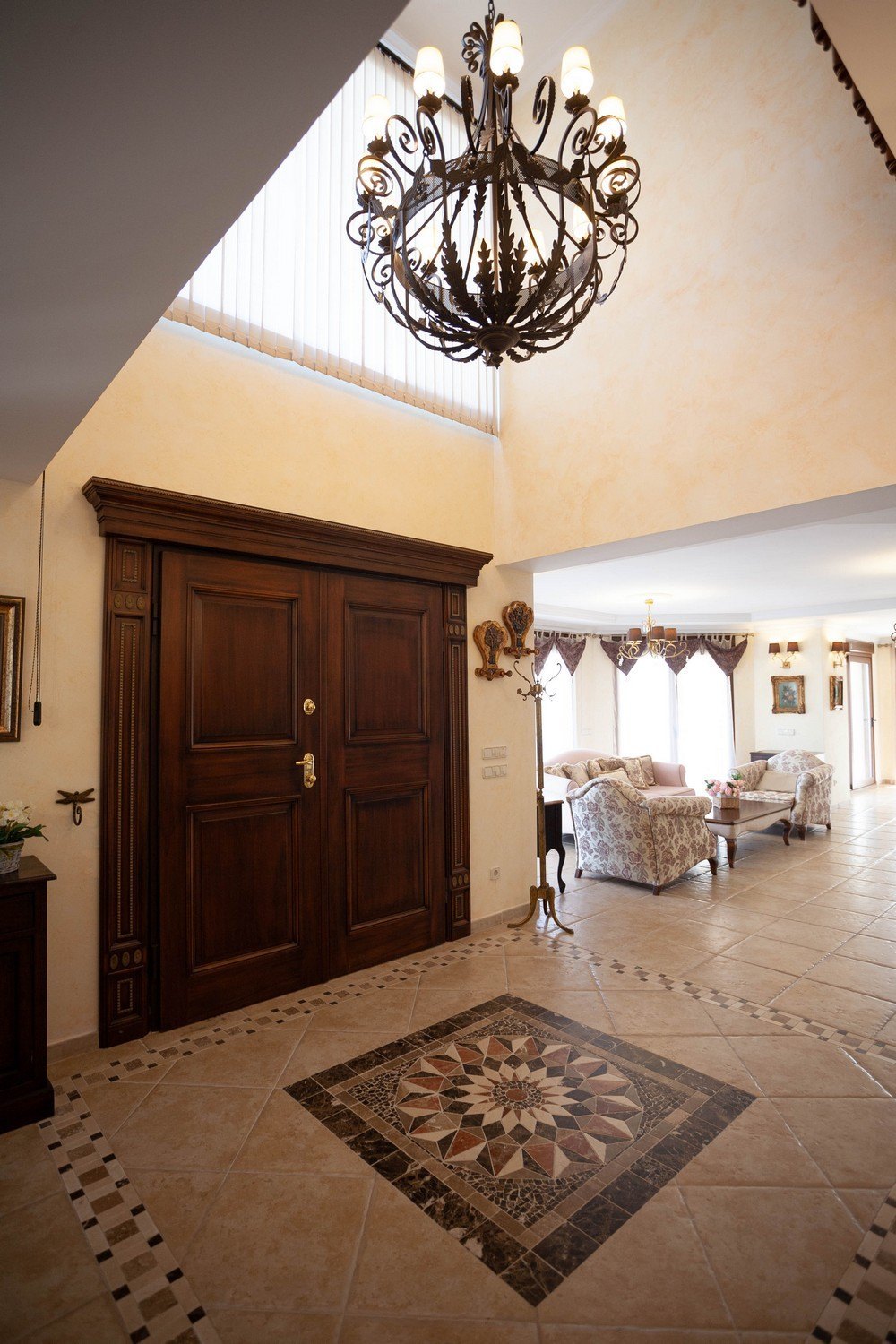 Vestíbulo con puerta doble de madera oscura y lámpara de araña ornamentada. Suelos de baldosas con diseño geométrico.