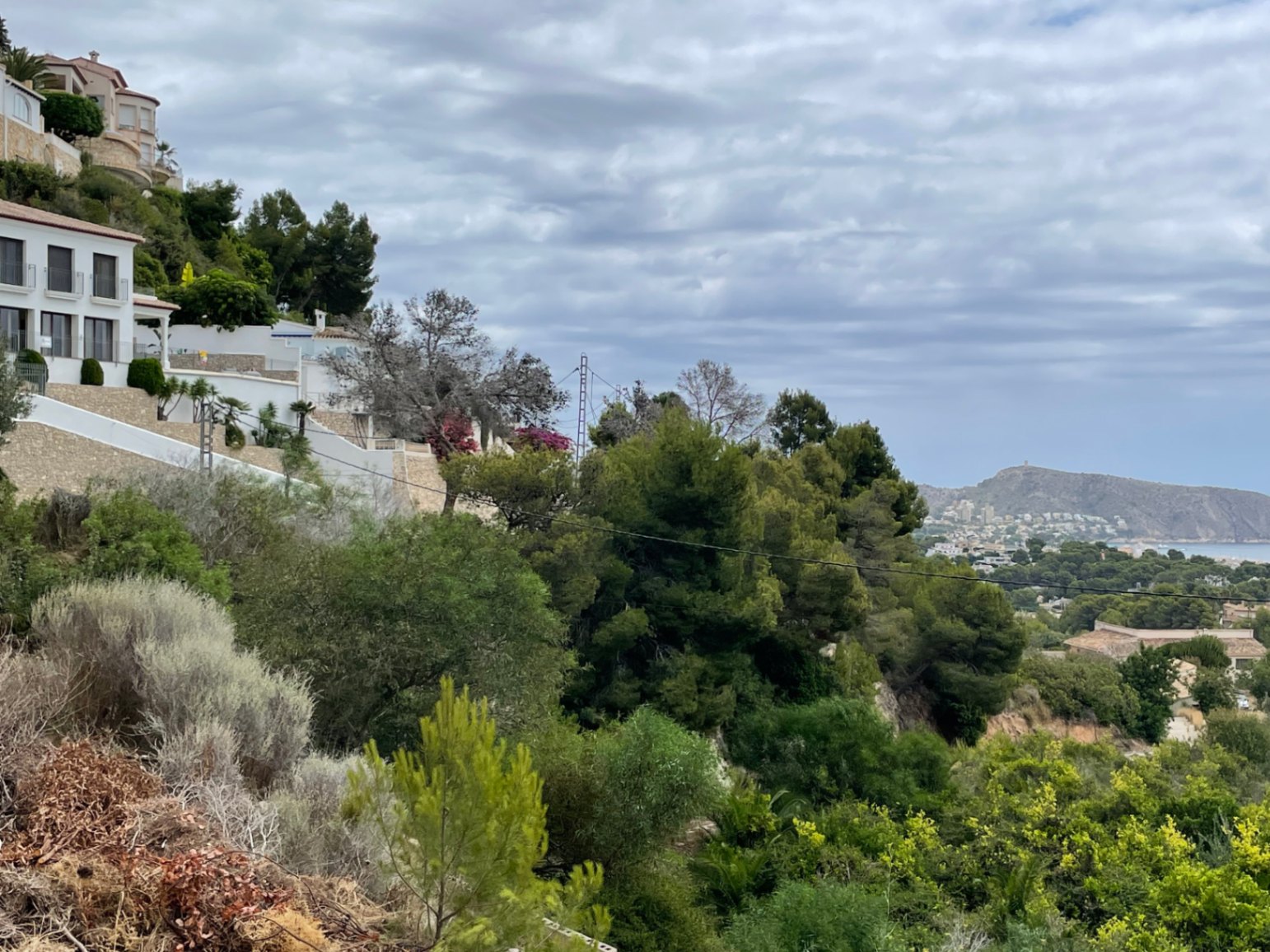 Vista elevada desde la parcela en Benimeit, destacando la vegetación mediterránea, muros de piedra y la silueta del Cap d'Or sobre el mar al fondo.
