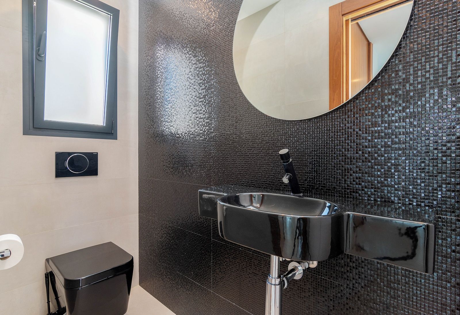 Aseo de cortesía con pared de mosaico negro, lavabo suspendido a juego y sanitario de pared. Incluye un espejo circular y ventana oscilobatiente.