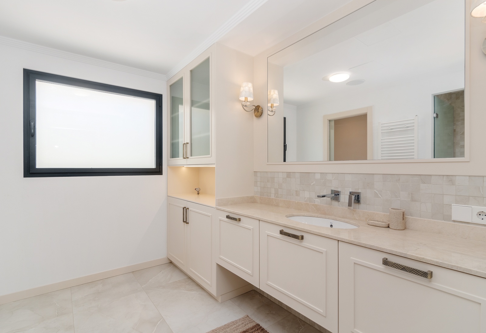 Baño elegante con encimera de piedra, lavabo integrado y mobiliario en tonos crema. Incluye armarios de vitrina con iluminación y apliques de pared clásicos junto al espejo.