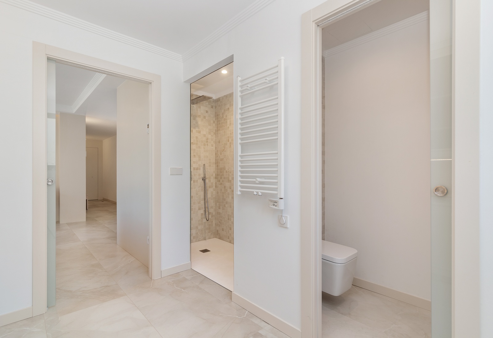 Baño moderno con ducha de obra en mosaico, inodoro suspendido, radiador toallero blanco y puertas correderas de cristal sobre suelos de gres porcelánico.