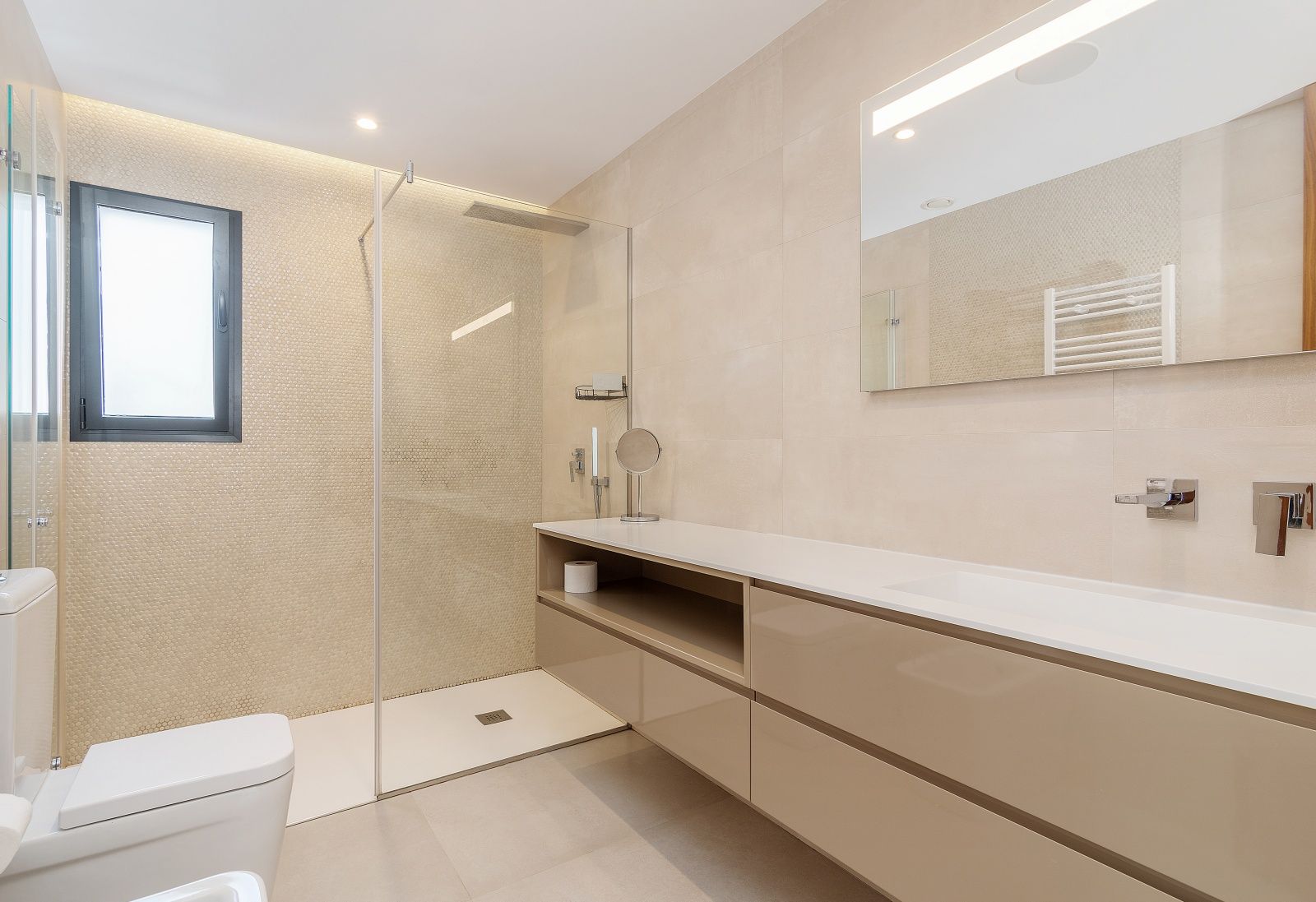 Baño moderno con ducha de obra, mampara de cristal, mueble suspendido con lavabo integrado y revestimientos en tonos beige con detalles de mosaico.