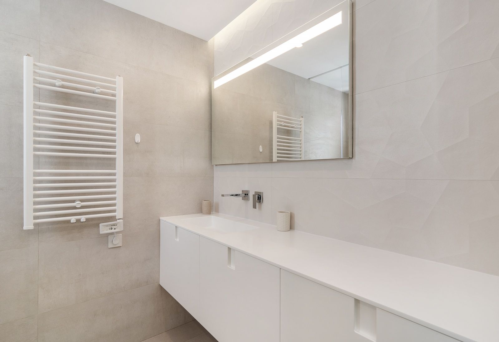Baño moderno con mueble suspendido blanco, grifería empotrada cromada, espejo con iluminación LED y radiador toallero sobre paredes de gres porcelánico con relieve.