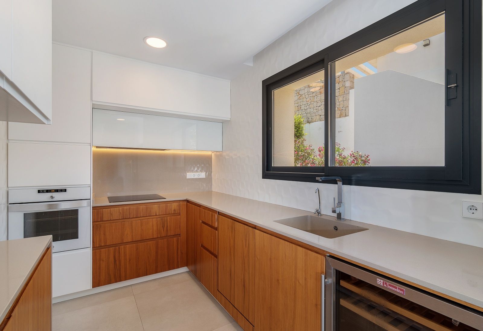 Cocina de diseño con armarios de madera, encimera de cuarzo, electrodomésticos integrados, vinoteca y ventana panorámica hacia el patio con muros de piedra.