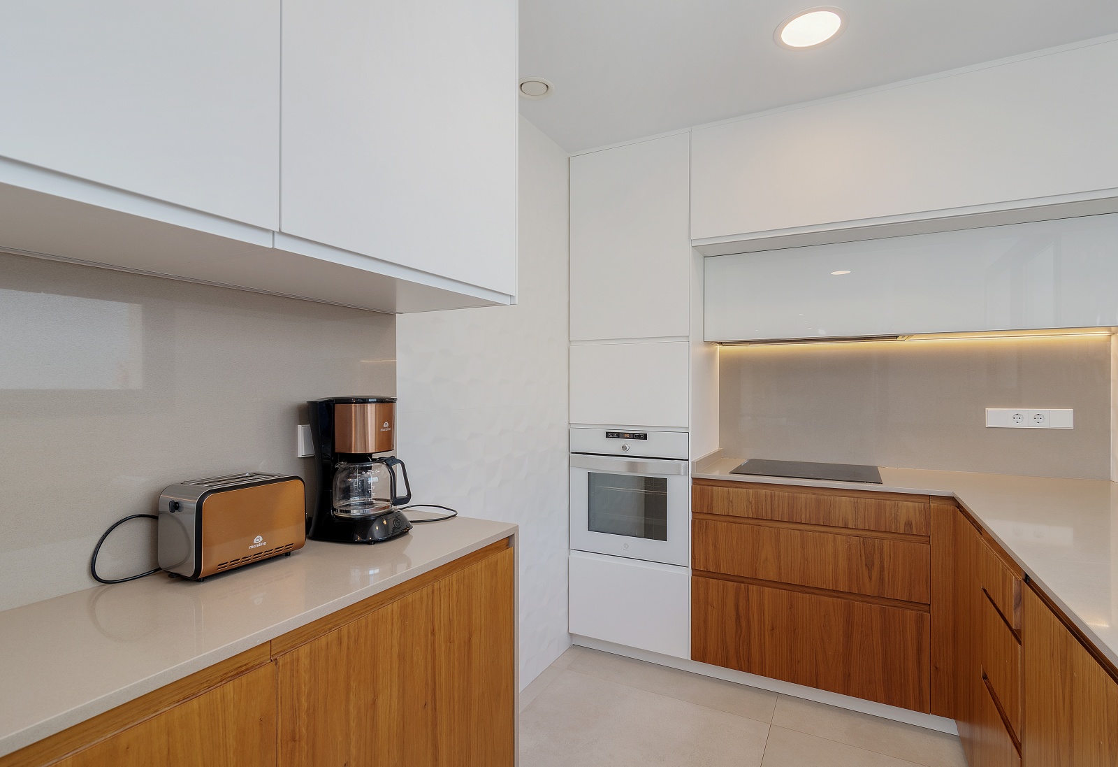 Cocina moderna con armarios de dos tonos en blanco y madera, encimera de cuarzo gris, horno integrado y placa de inducción con iluminación LED bajo los muebles.
