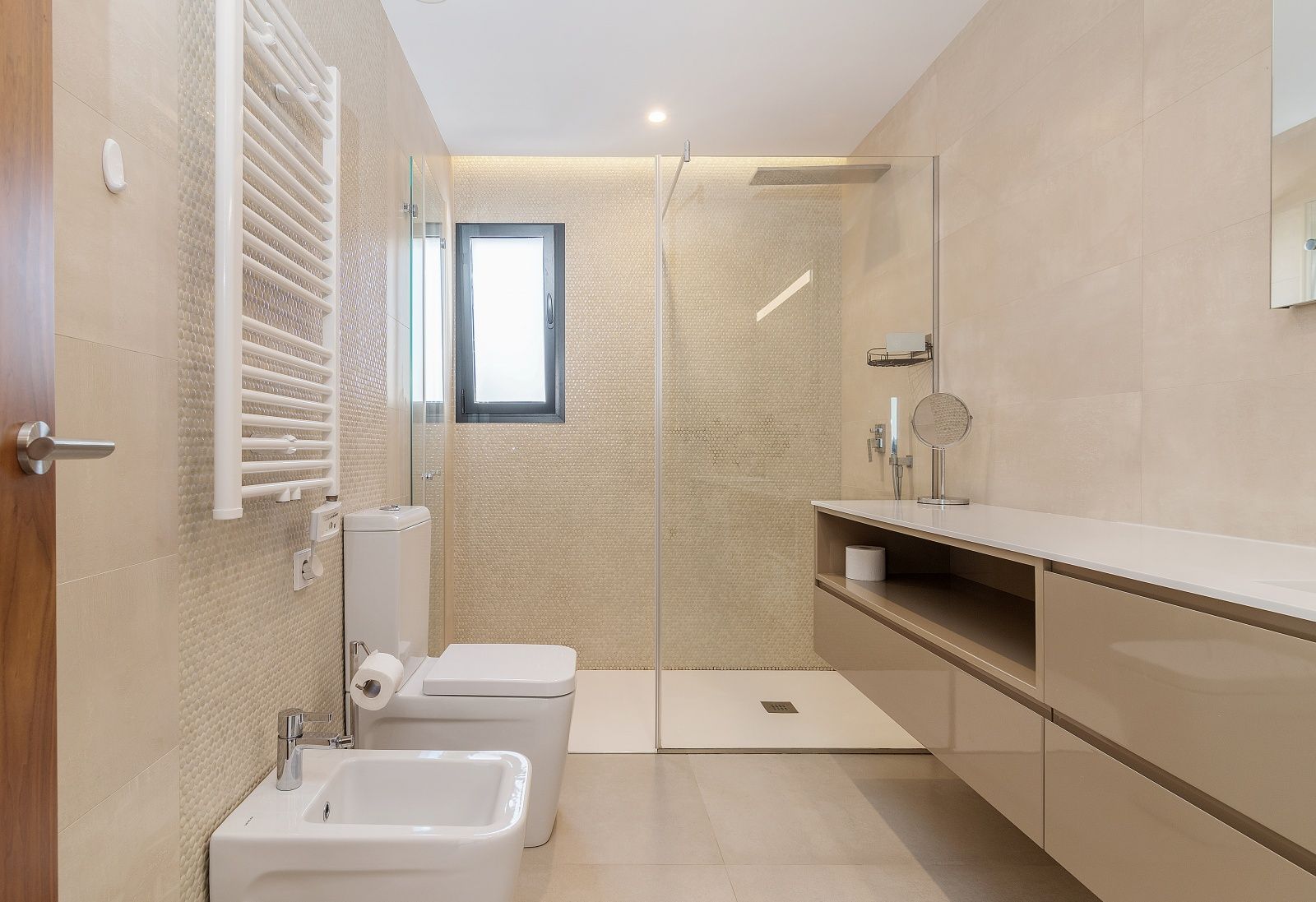 Cuarto de baño moderno con ducha a ras de suelo, mampara de cristal, mueble suspendido, bidé y radiador toallero blanco sobre gres cerámico beige.