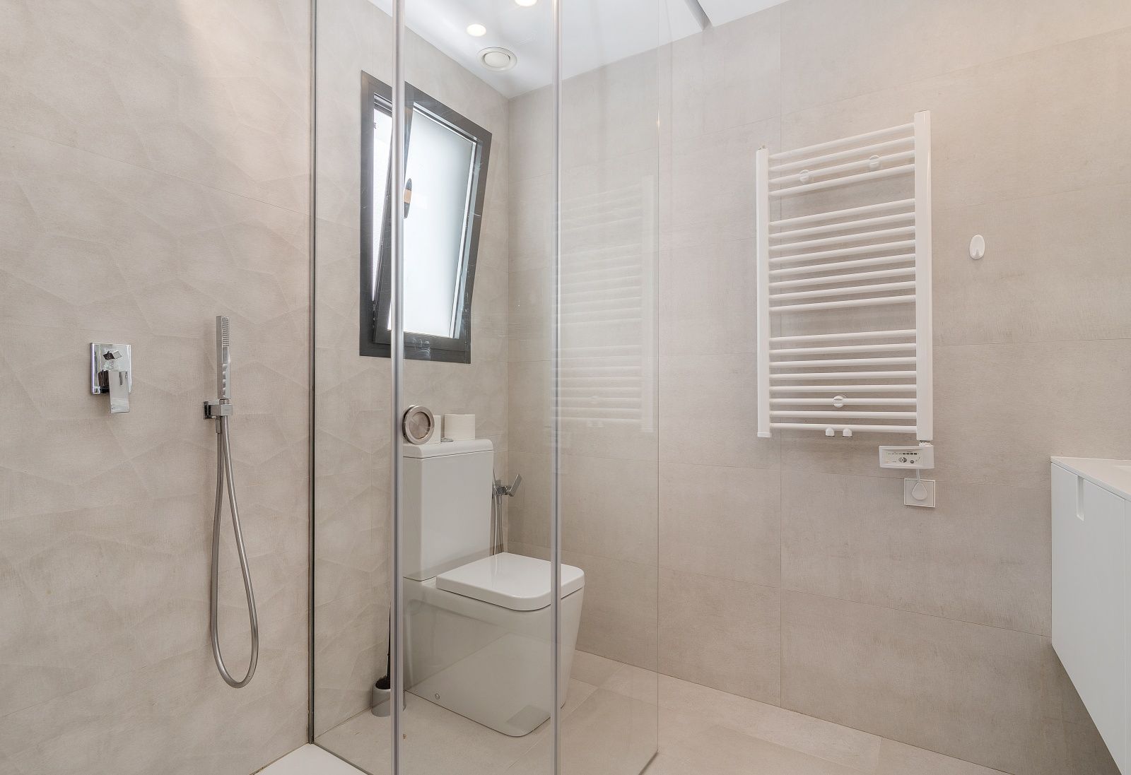 Cuarto de baño moderno con ducha de obra acristalada, revestimiento cerámico texturizado en tonos beige y radiador toallero blanco de pared.