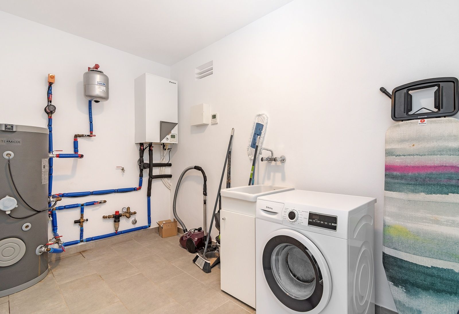 Cuarto de lavado y sala técnica equipada con lavadora, fregadero de servicio e instalaciones visibles de aerotermia y fontanería sobre suelo cerámico.
