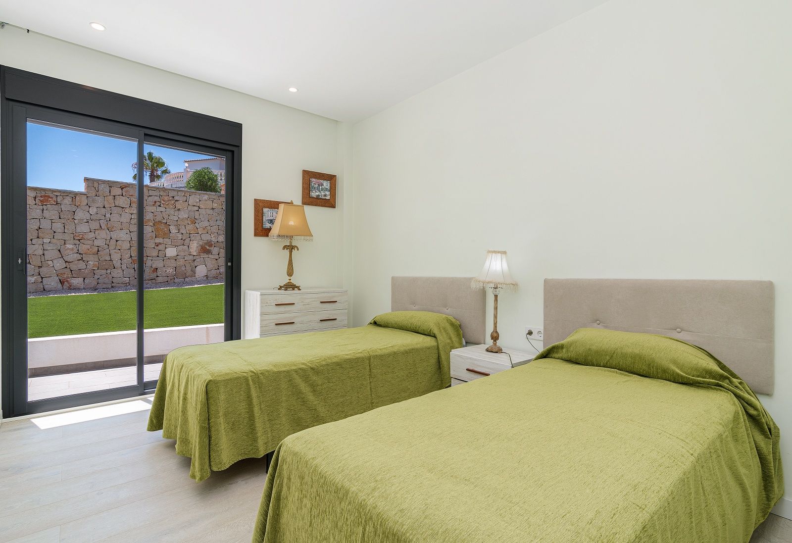 Dormitorio con dos camas individuales, cabeceros tapizados y acceso directo al jardín mediante ventanal corredero. Suelos de madera clara y paredes en tonos neutros.