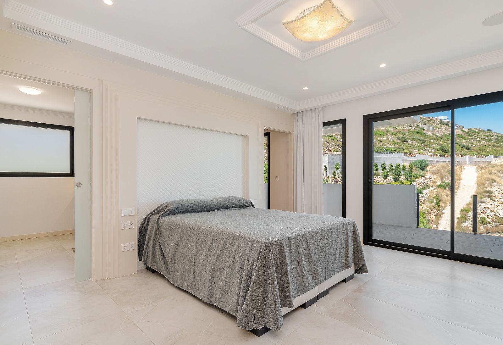 Dormitorio luminoso con acceso directo a la terraza mediante ventanales correderos, molduras decorativas en el techo y cabecero integrado en la pared.