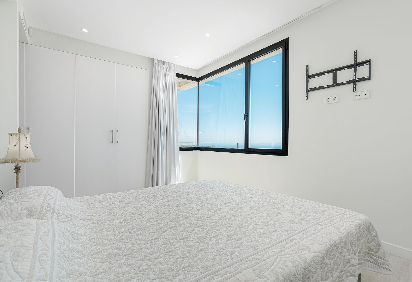 Dormitorio luminoso con armarios empotrados blancos, iluminación empotrada y ventanal de esquina con marcos negros que ofrece vistas despejadas al mar Mediterráneo.
