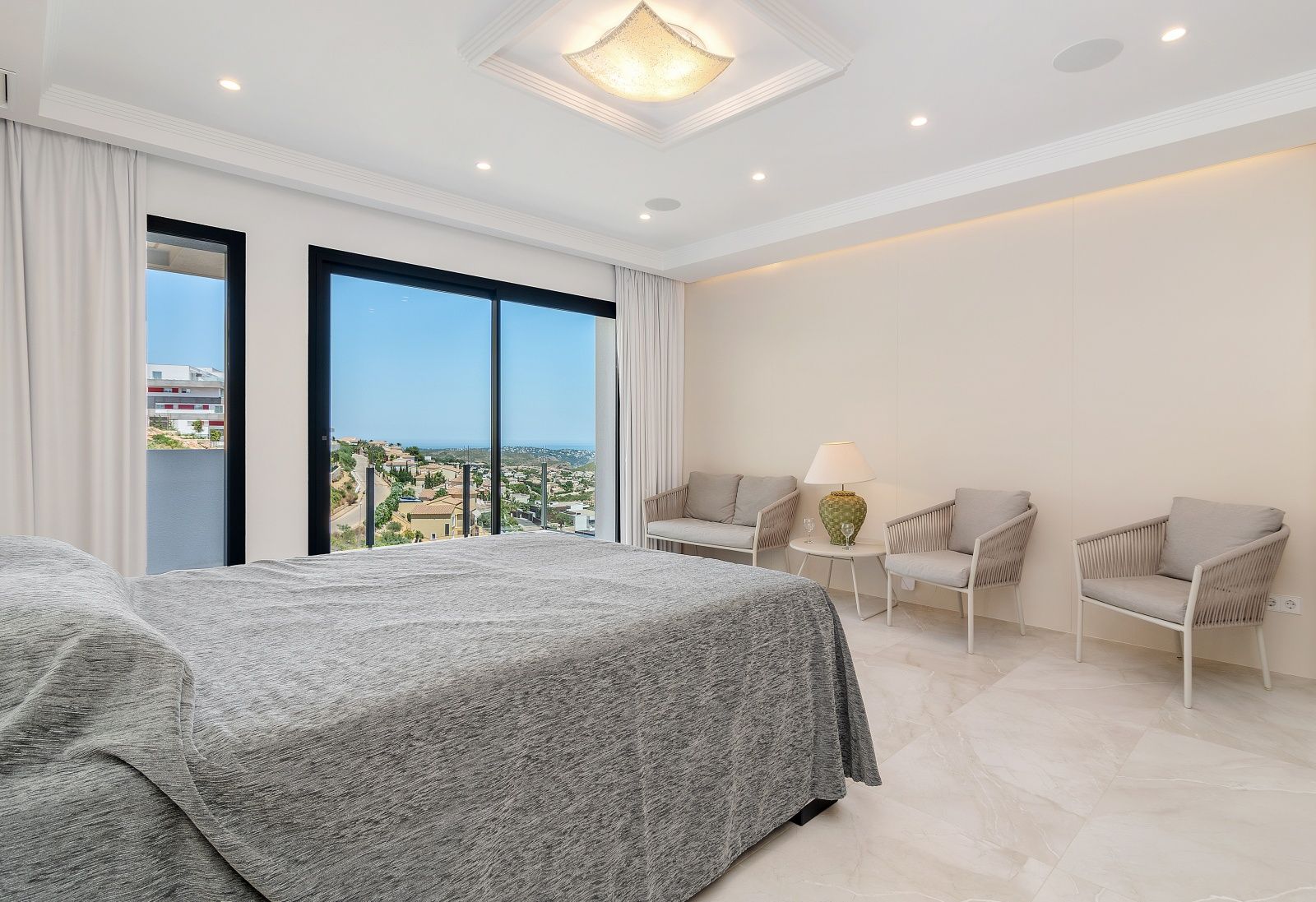 Dormitorio luminoso con suelos de gres porcelánico, zona de estar integrada y grandes ventanales con vistas panorámicas al mar y a la urbanización Cumbre del Sol.