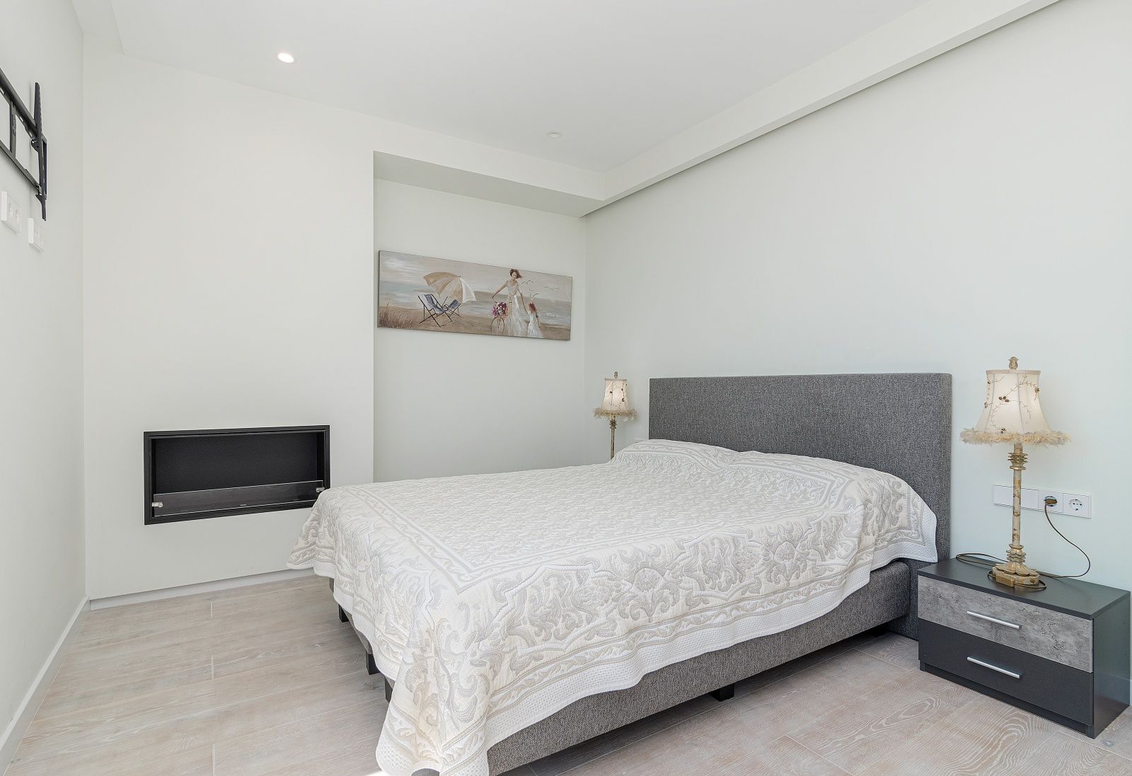 Dormitorio moderno con chimenea de pared integrada, suelos de gres porcelánico efecto madera y cama con cabecero tapizado en gris.